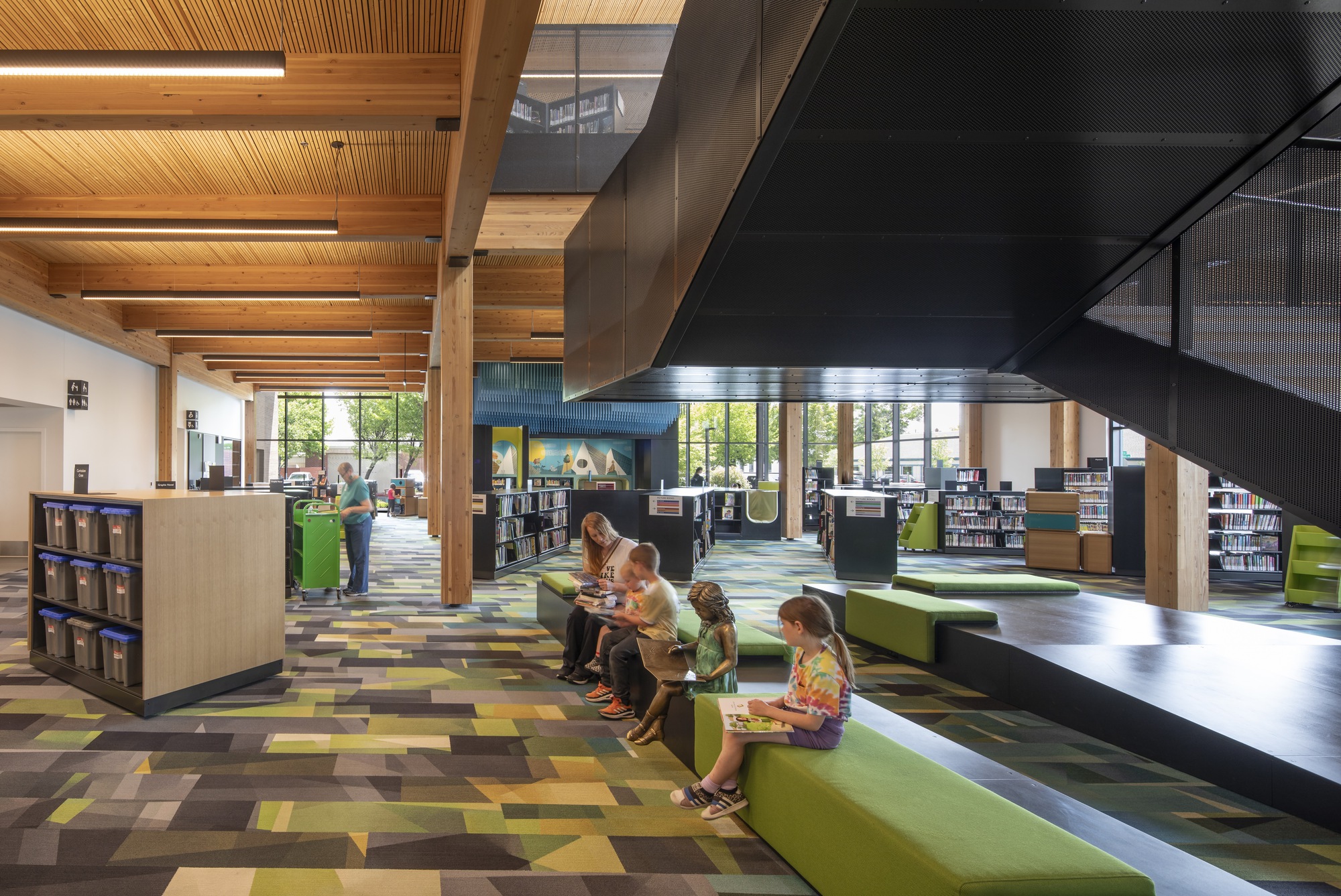 Library reading area with green stepped seating platform beneath a suspended metal staircase and exposed timber ceiling