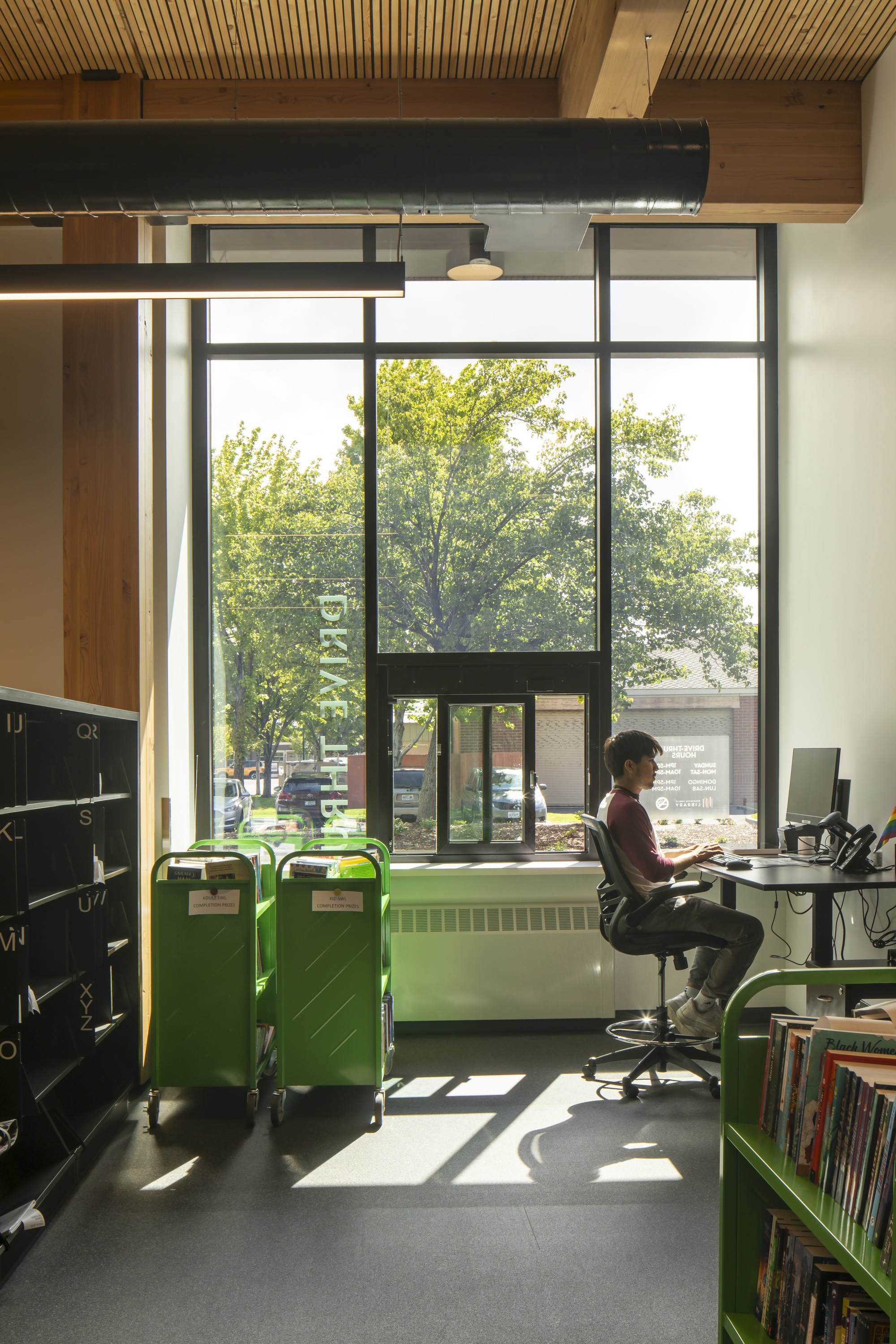Workstation beneath a steel beam and timber ceiling with green bins and tall windows overlooking street trees