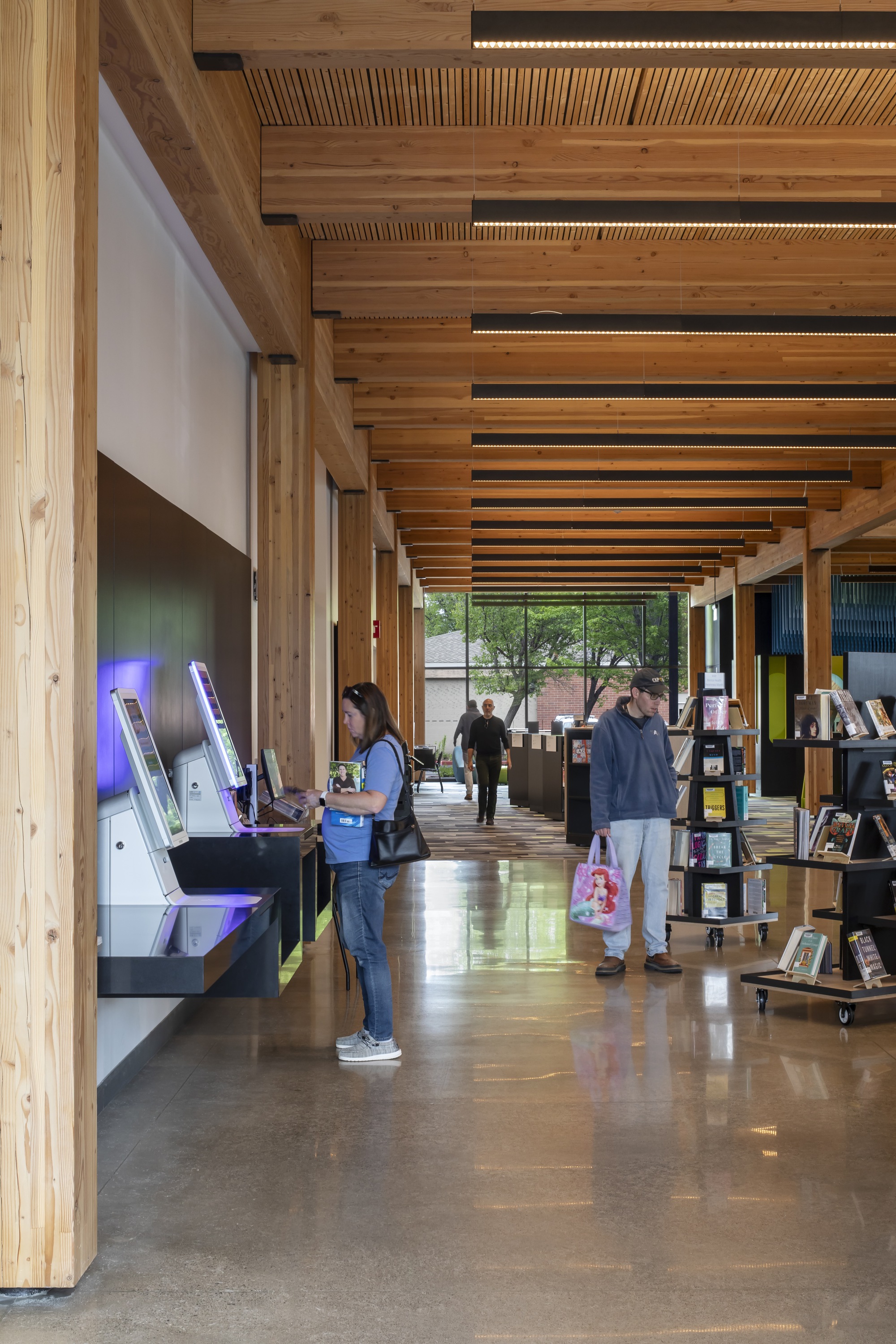 Long library corridor with rhythmic timber beams and ceiling slats as patrons use digital catalog stations
