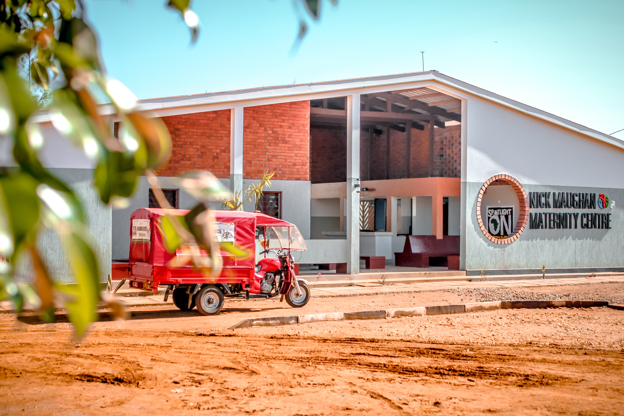 Street view of the white-rendered facade with exposed brick panels and a red three-wheeled vehicle on the dirt road
