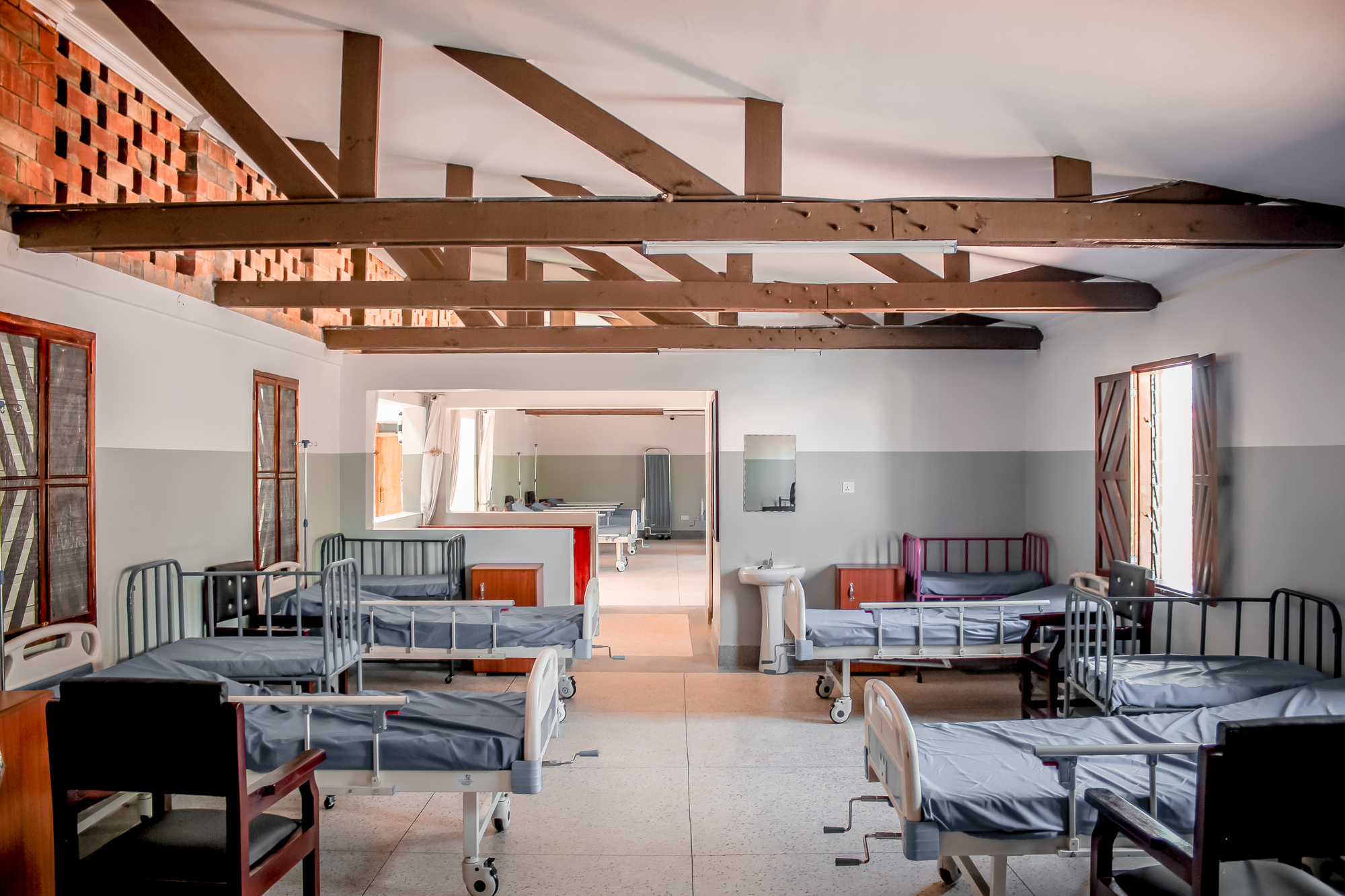 Interior ward with exposed timber trusses, hospital beds and a perforated brick screen visible through the doorway