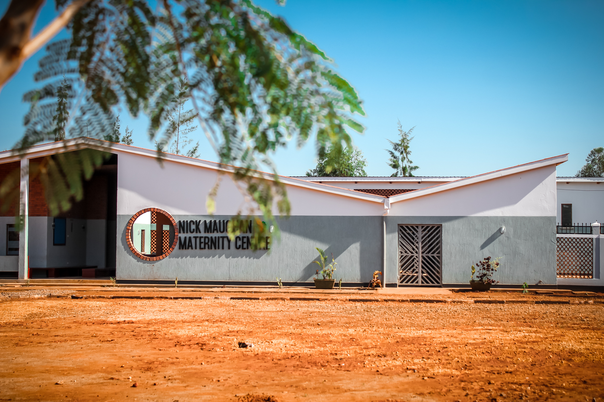 Front facade with butterfly roof and circular logo seen through overhanging foliage in midday sun