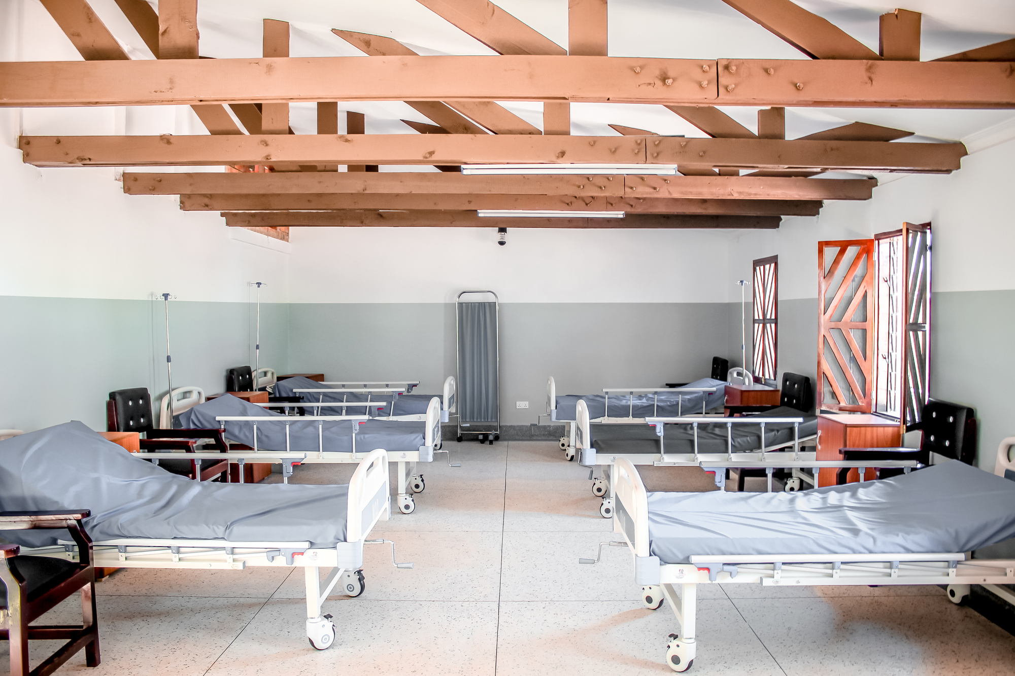 Interior ward with metal hospital beds beneath an exposed timber beam ceiling and grey wainscoting