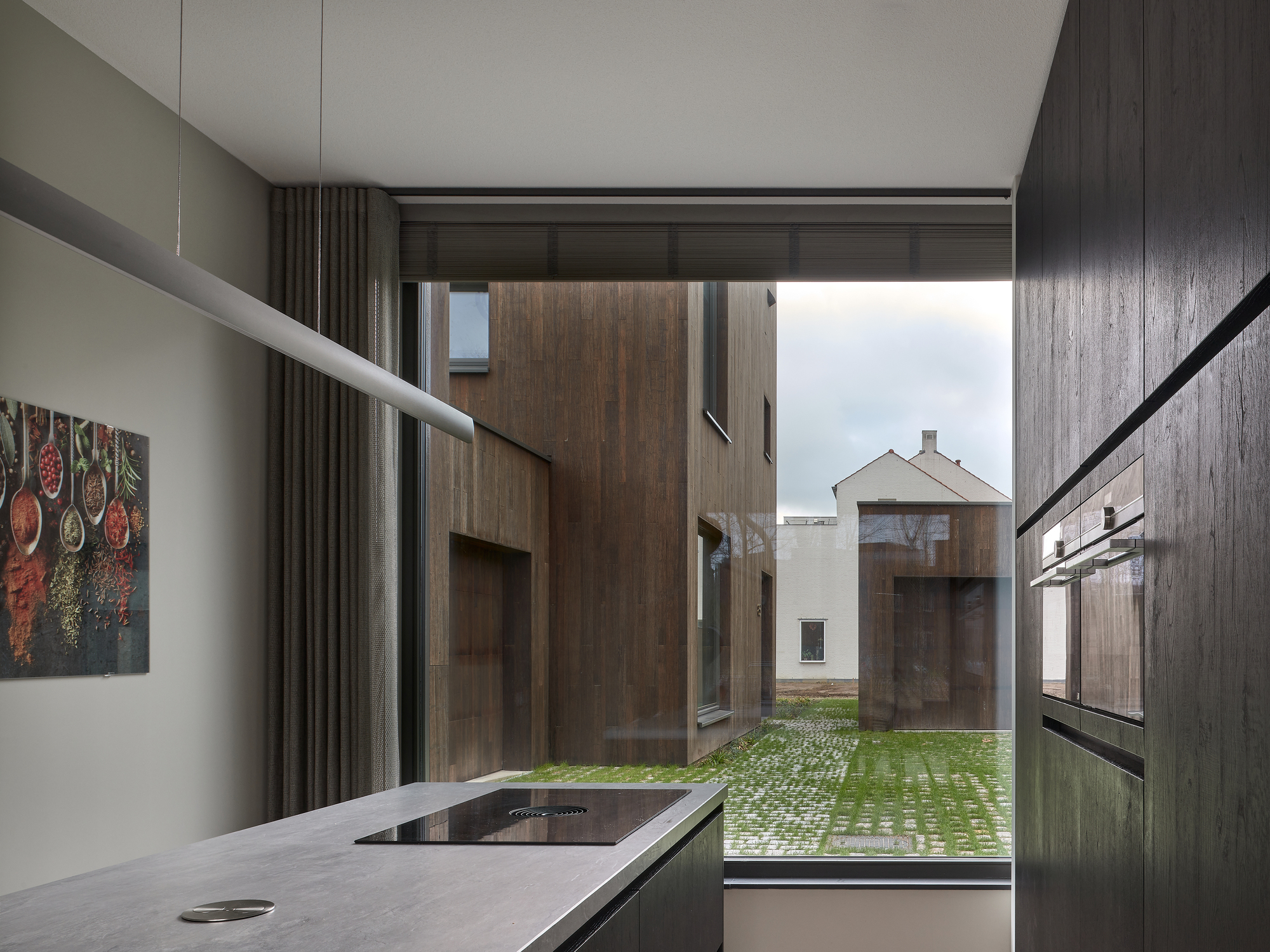 Kitchen window framing a view to timber-clad volumes across a courtyard with permeable paving