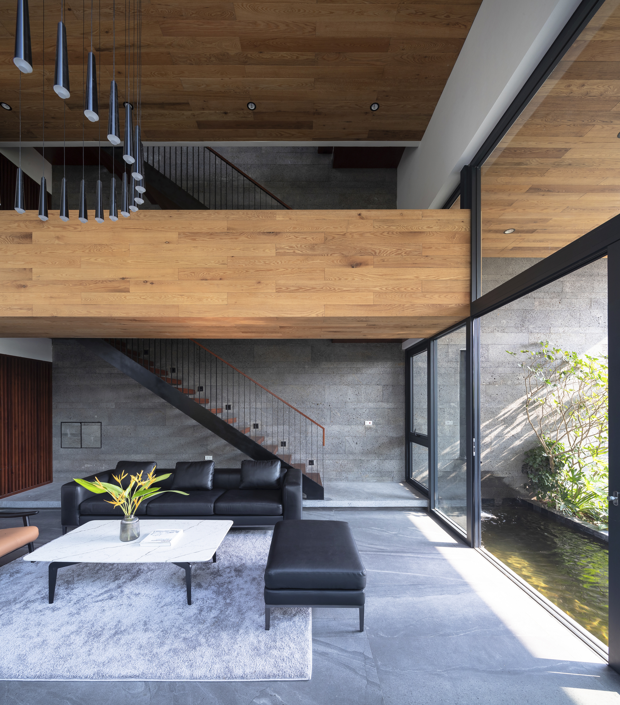 Double-height living room with timber bridge, concrete staircase and glazed wall overlooking interior courtyard pond
