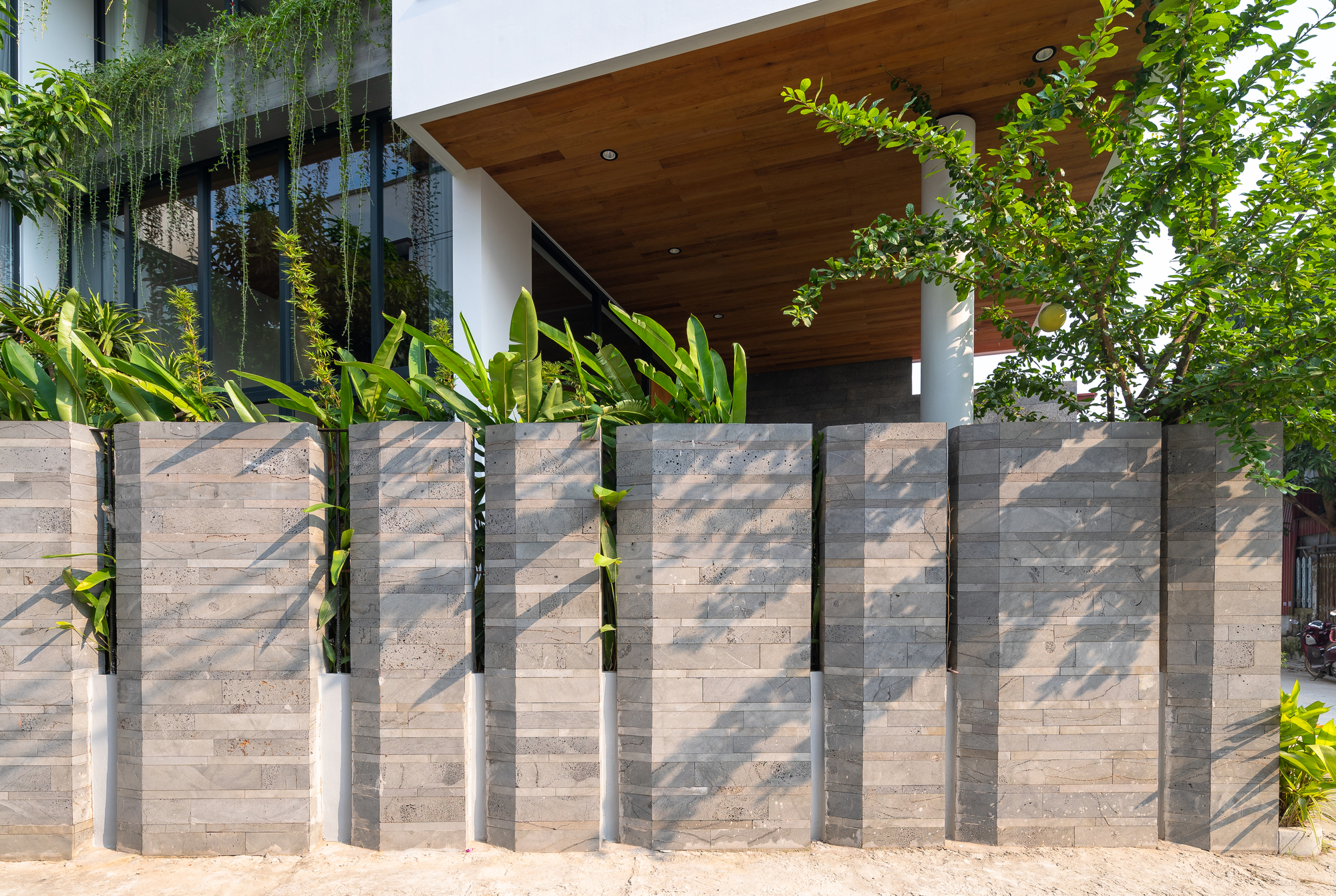 Layered concrete fence with vertical steel fins casting shadows beside planted beds and timber entry canopy