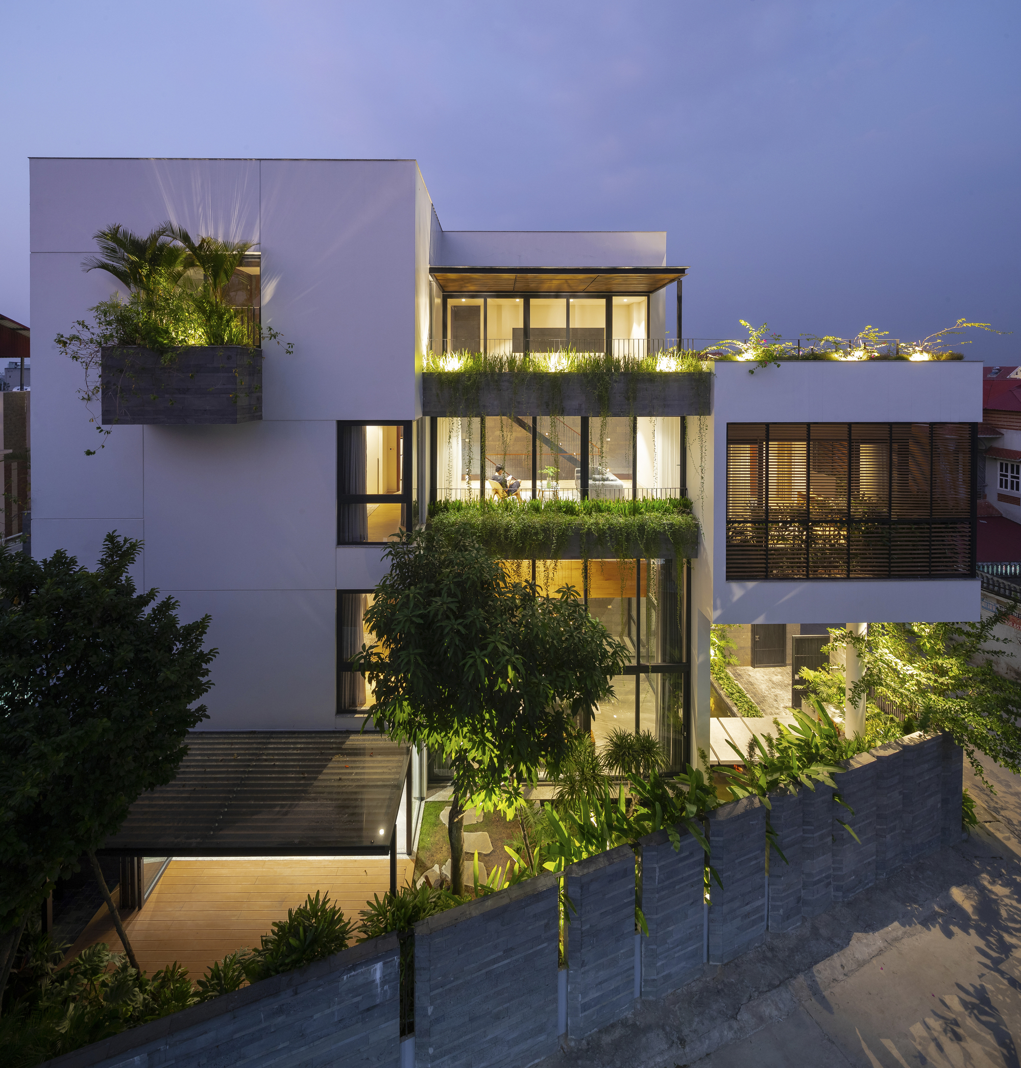 Street facade at twilight showing stacked planted terraces with uplighting and timber screening