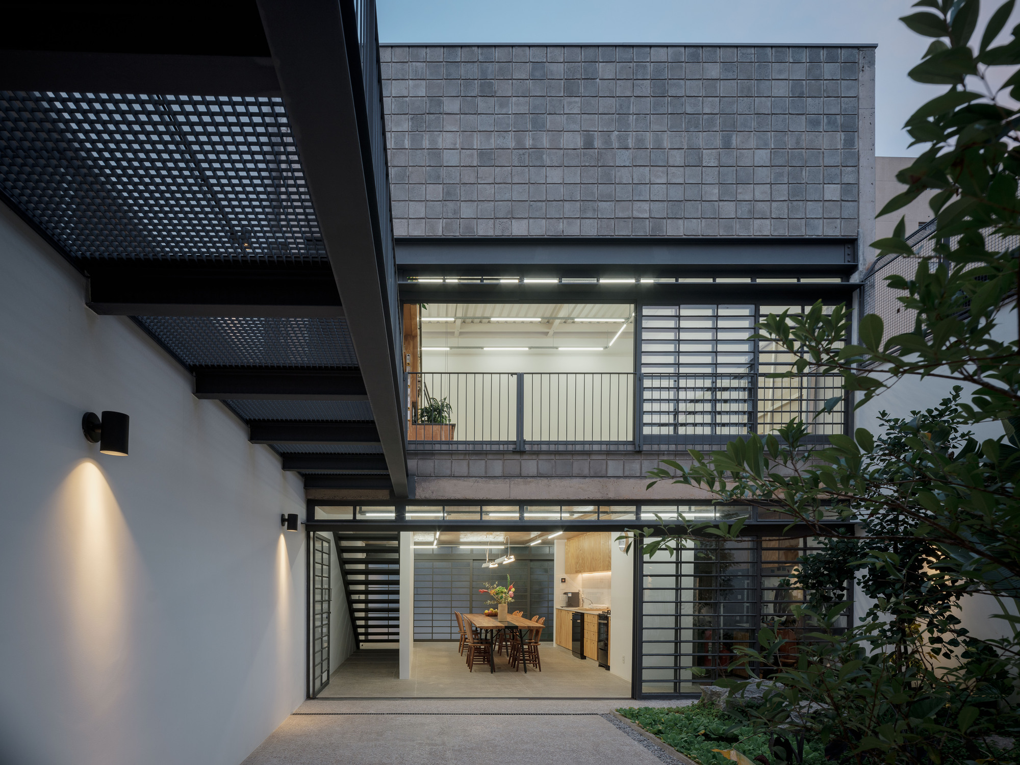 Courtyard facade at dusk with gray tile cladding, gridded screens, perforated metal stairs, and planted garden