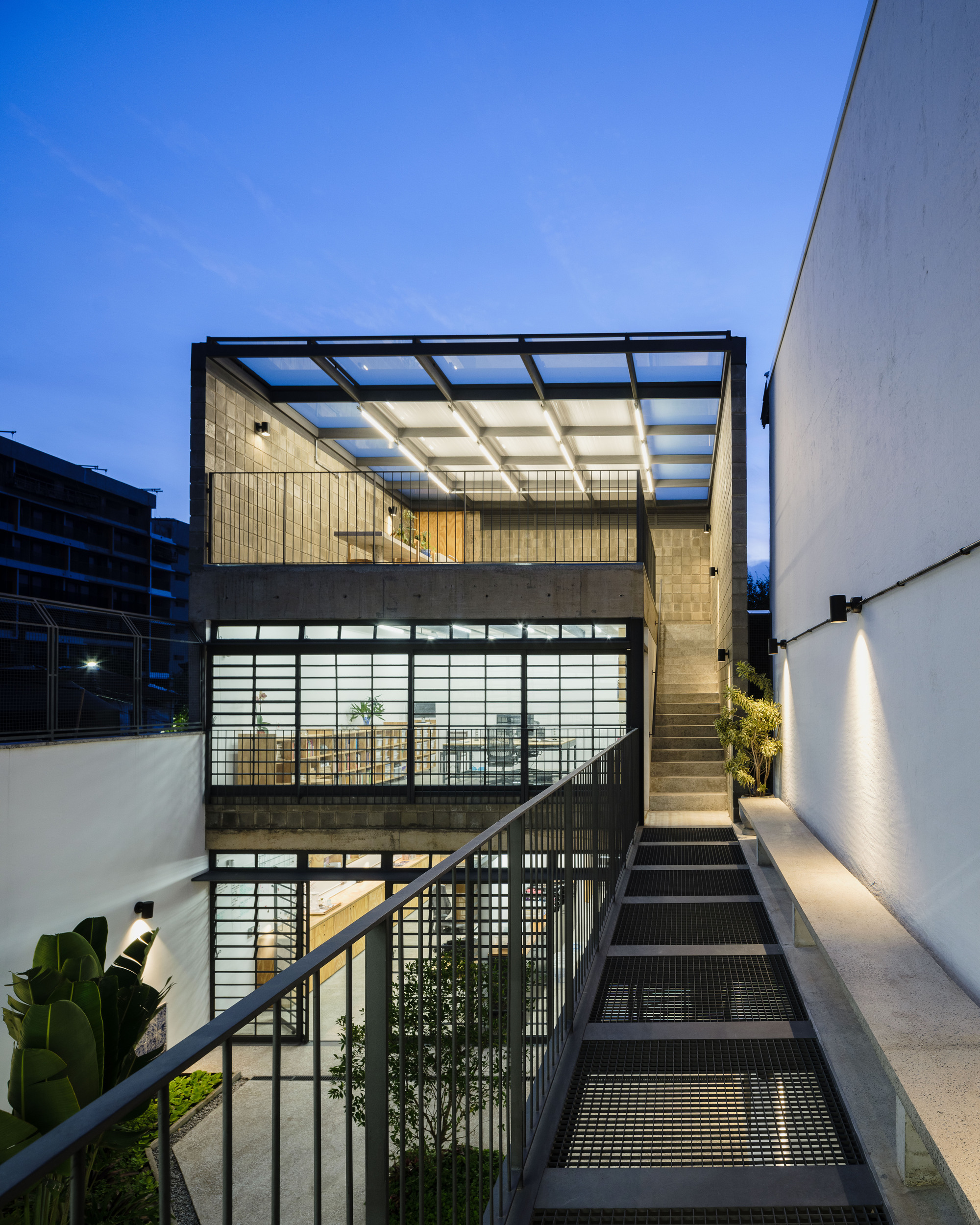 Exterior metal staircase ascending past translucent glazed walls and a glazed roof canopy at dusk