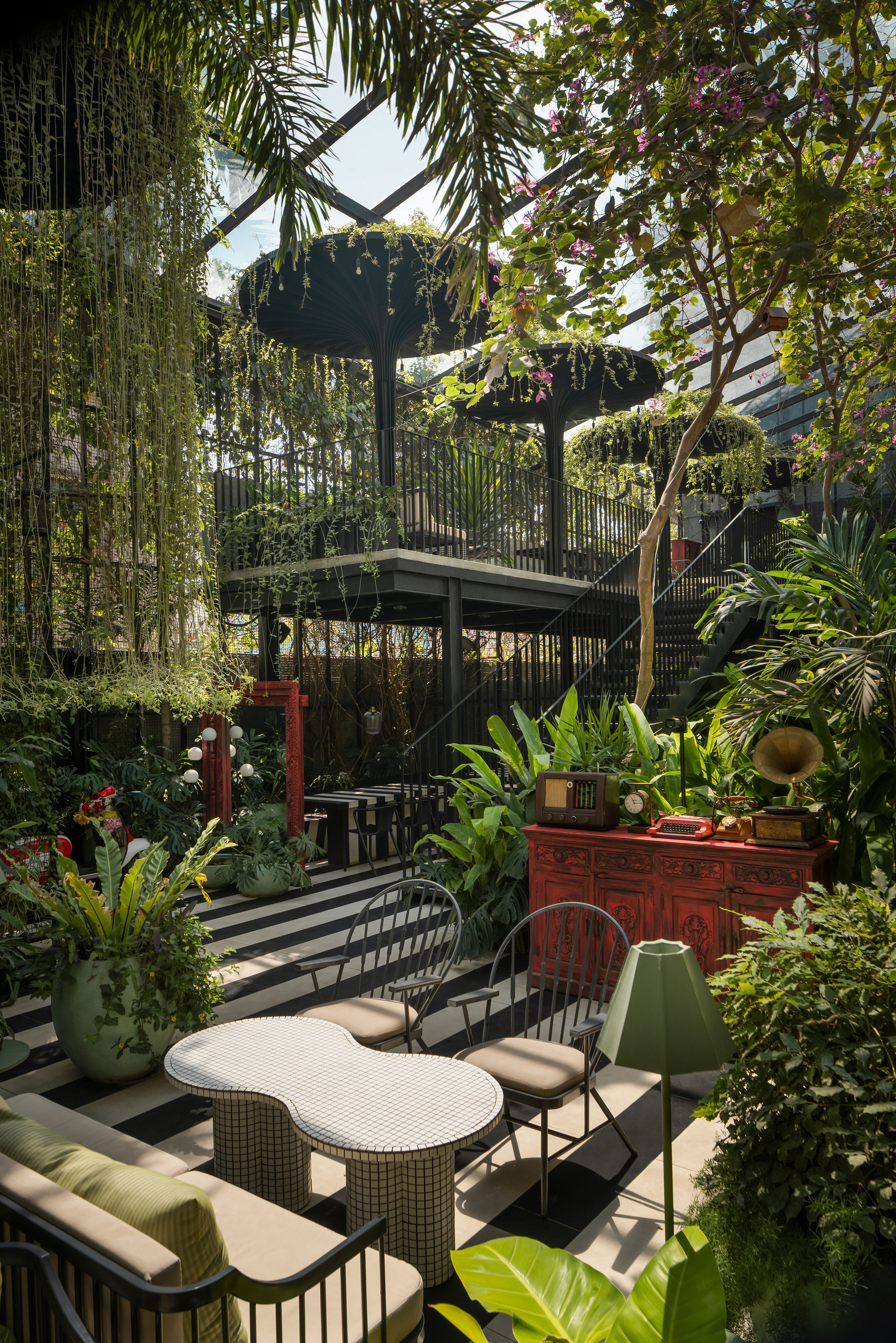 Outdoor seating area beneath a planted steel trellis system with hanging vines and tropical foliage