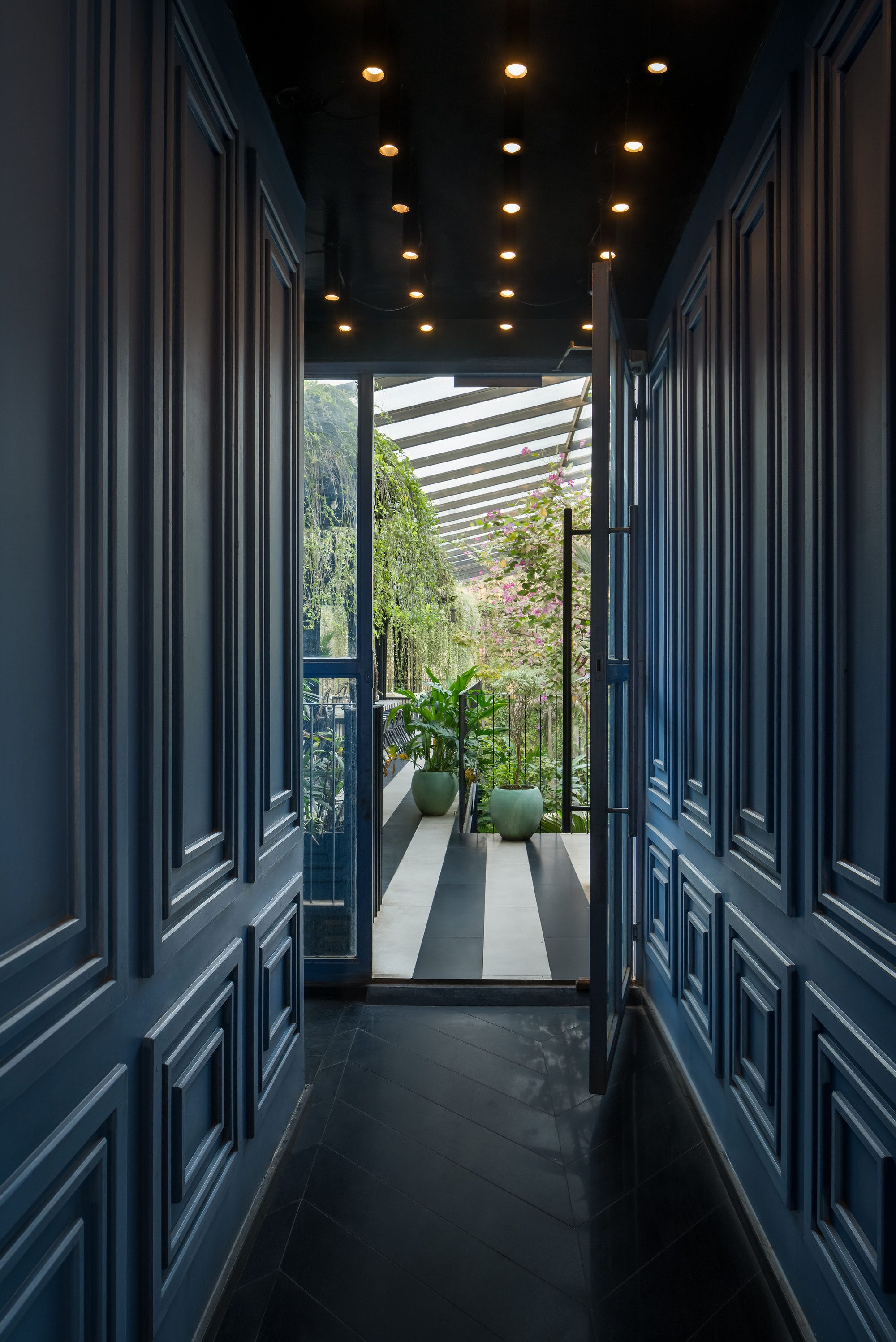 Narrow corridor with navy paneled walls and ceiling-mounted downlights leading to a planted terrace with slatted pergola