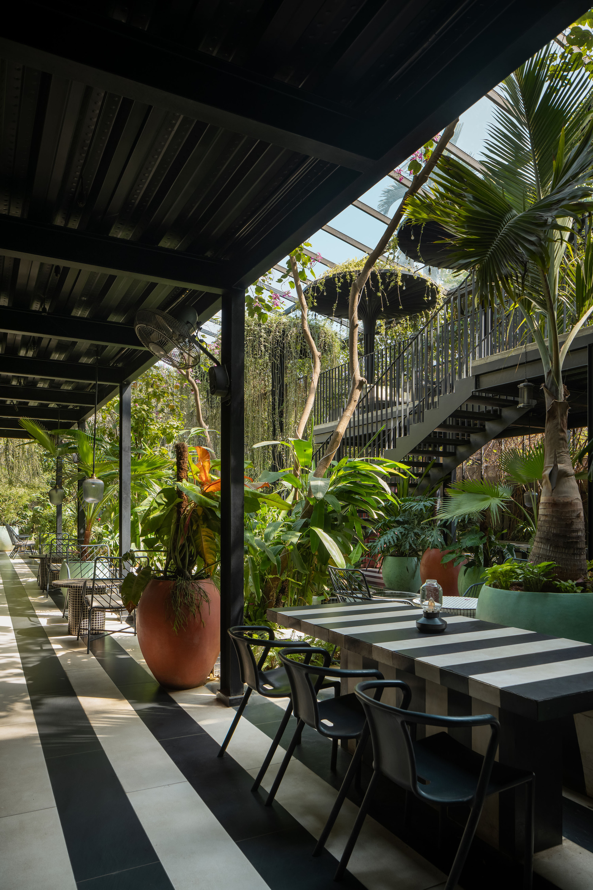 Covered dining terrace with exposed timber beams and striped floor tiles adjacent to garden with palms and hanging plants