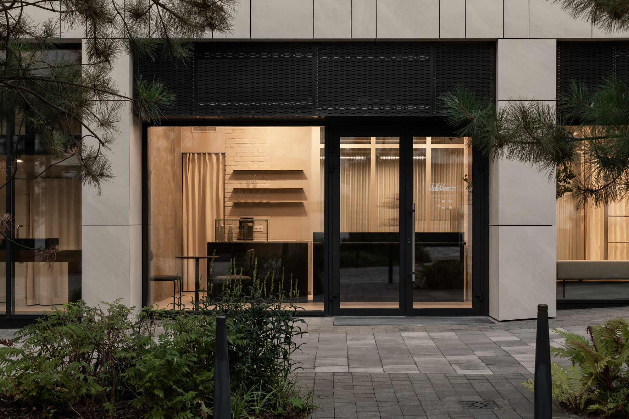 Street-level facade with black metal frames, timber interior millwork illuminated at dusk, and planted entry plaza