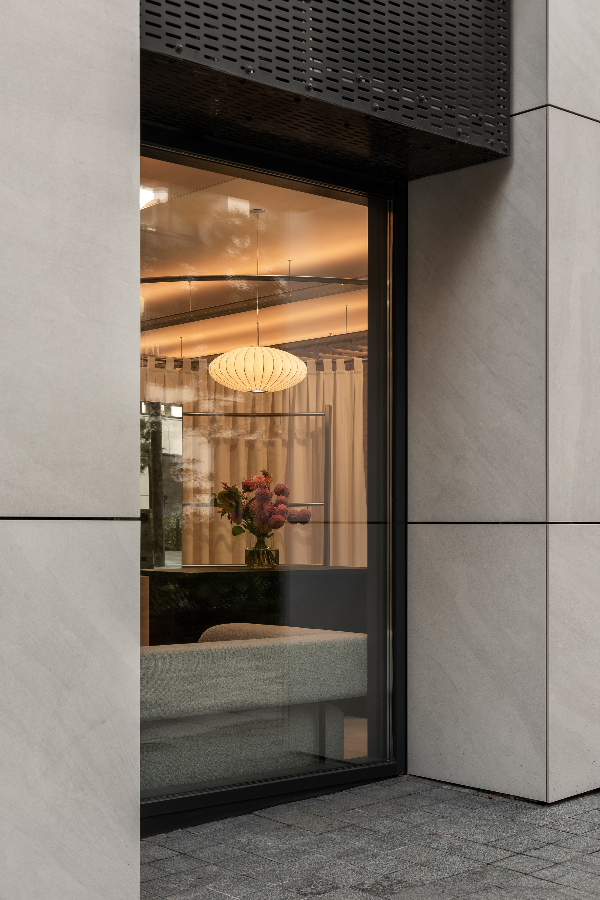 Corner window at twilight revealing interior with pleated pendant lamp and flowers reflected in glass