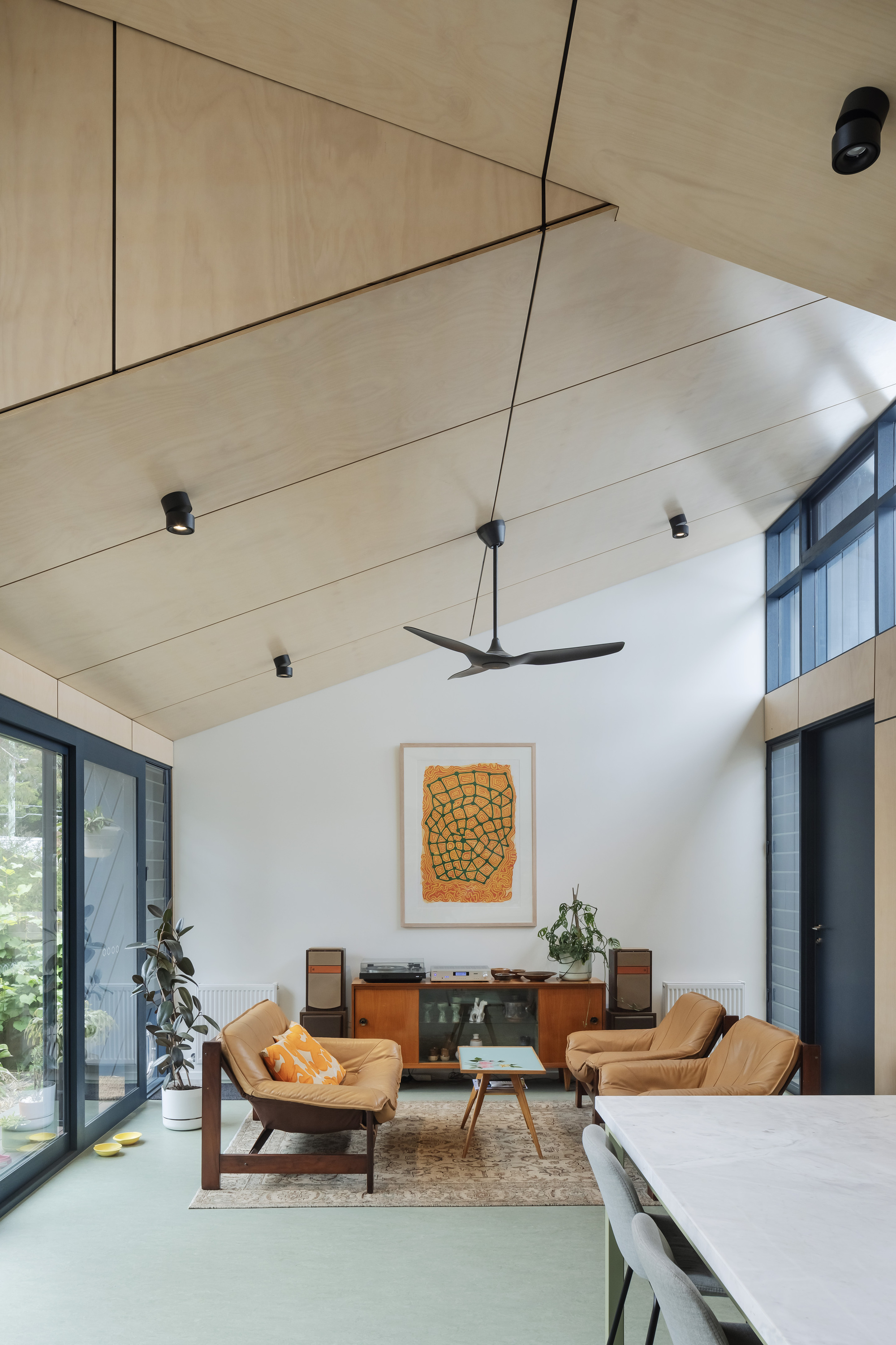 This architectural image showcases a modern living room with a vaulted plywood ceiling and abundant natural light. The space features mid-century modern furnitu