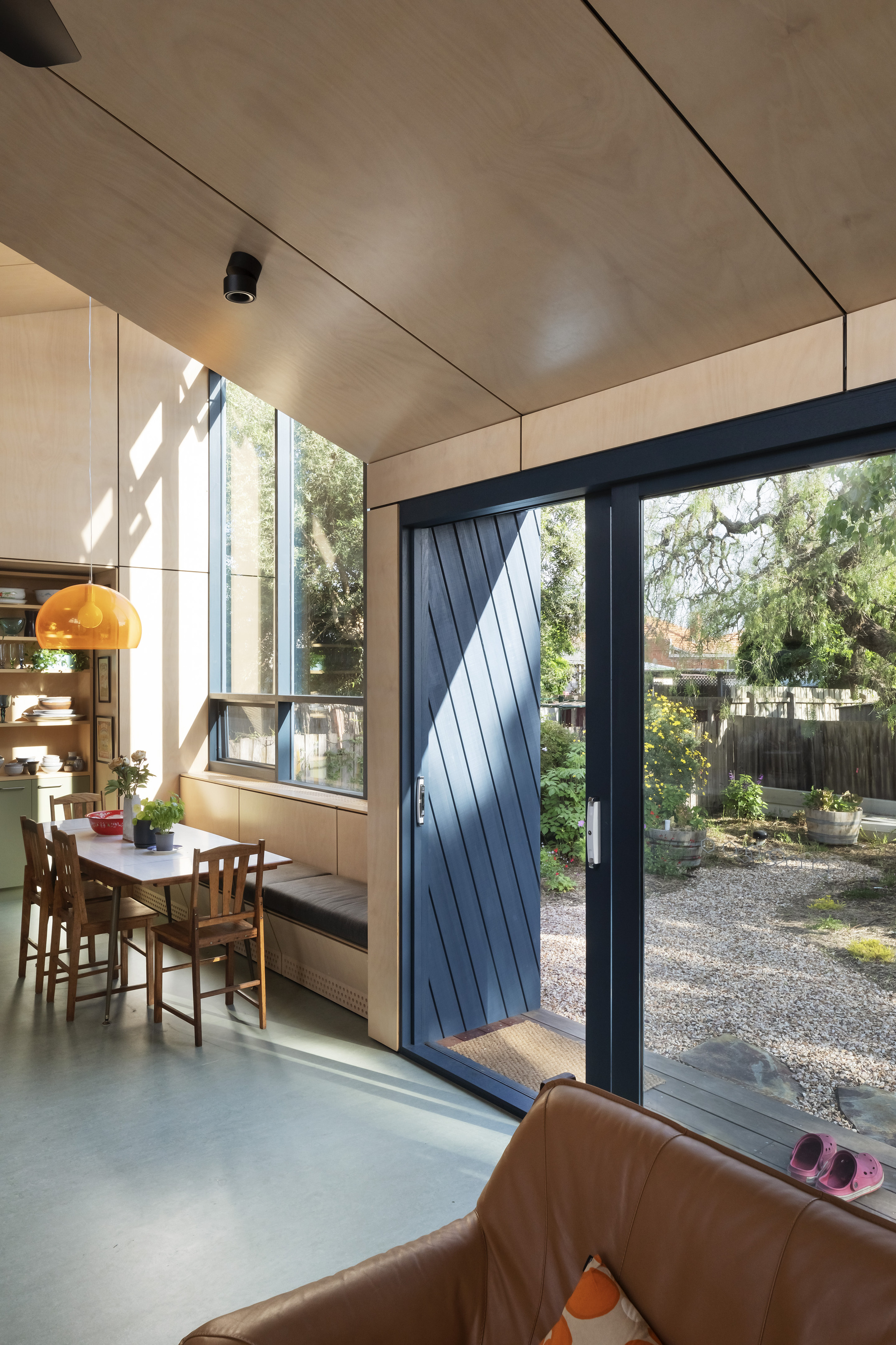 This architectural image showcases a modern interior with a focus on natural light and indoor-outdoor connection. The space features plywood paneling, a dining
