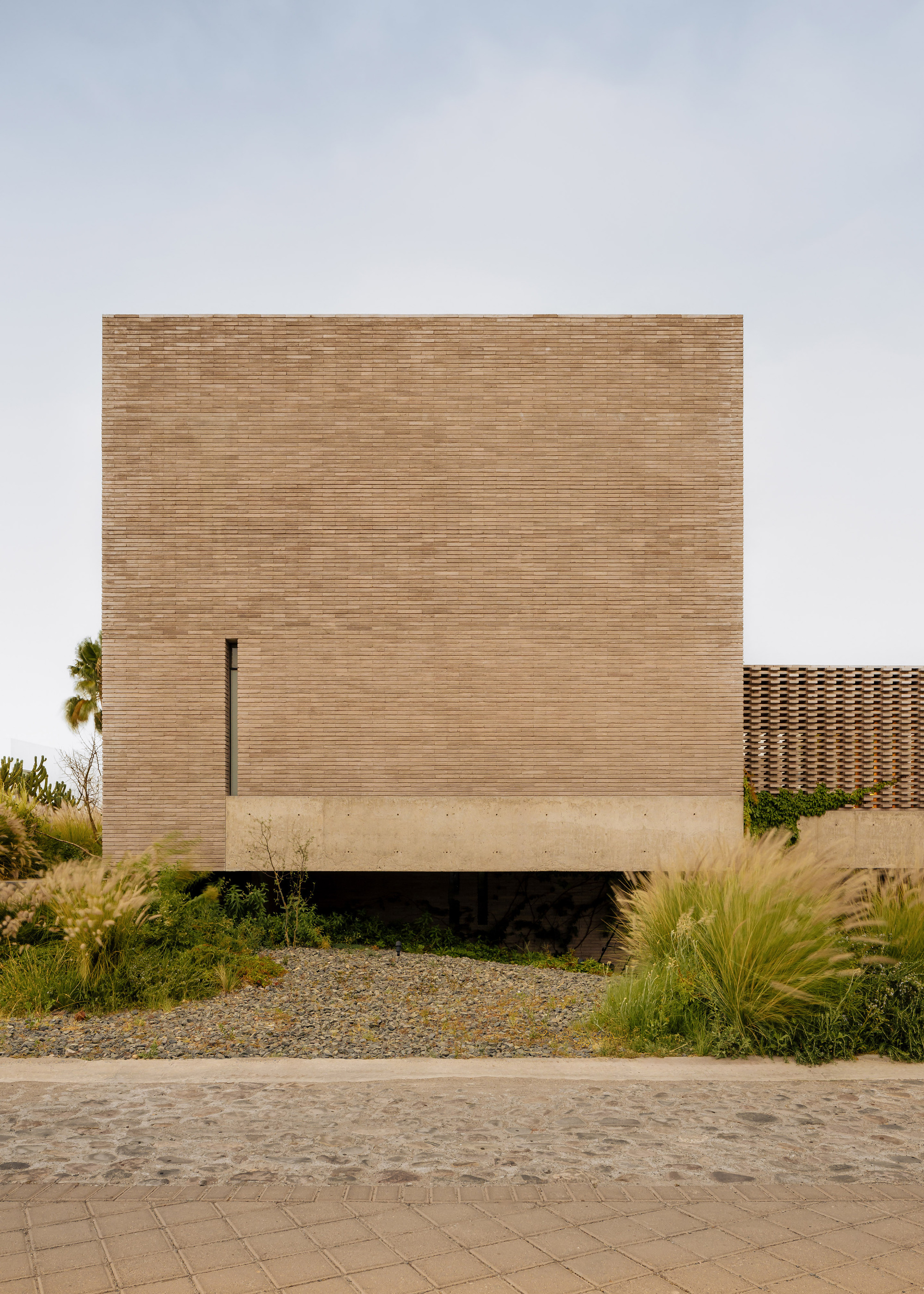 Front facade of horizontal brick volume with narrow vertical window slot and planted landscape bed