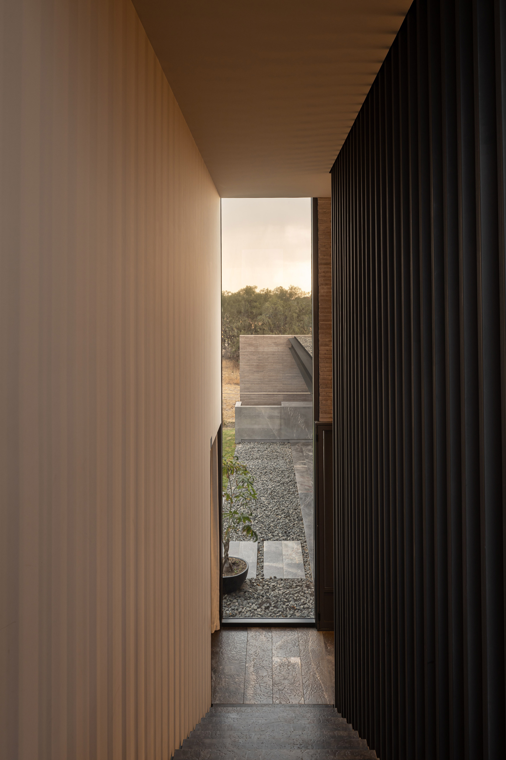 Vertical slit window between timber and black slat walls opening to a gravel courtyard