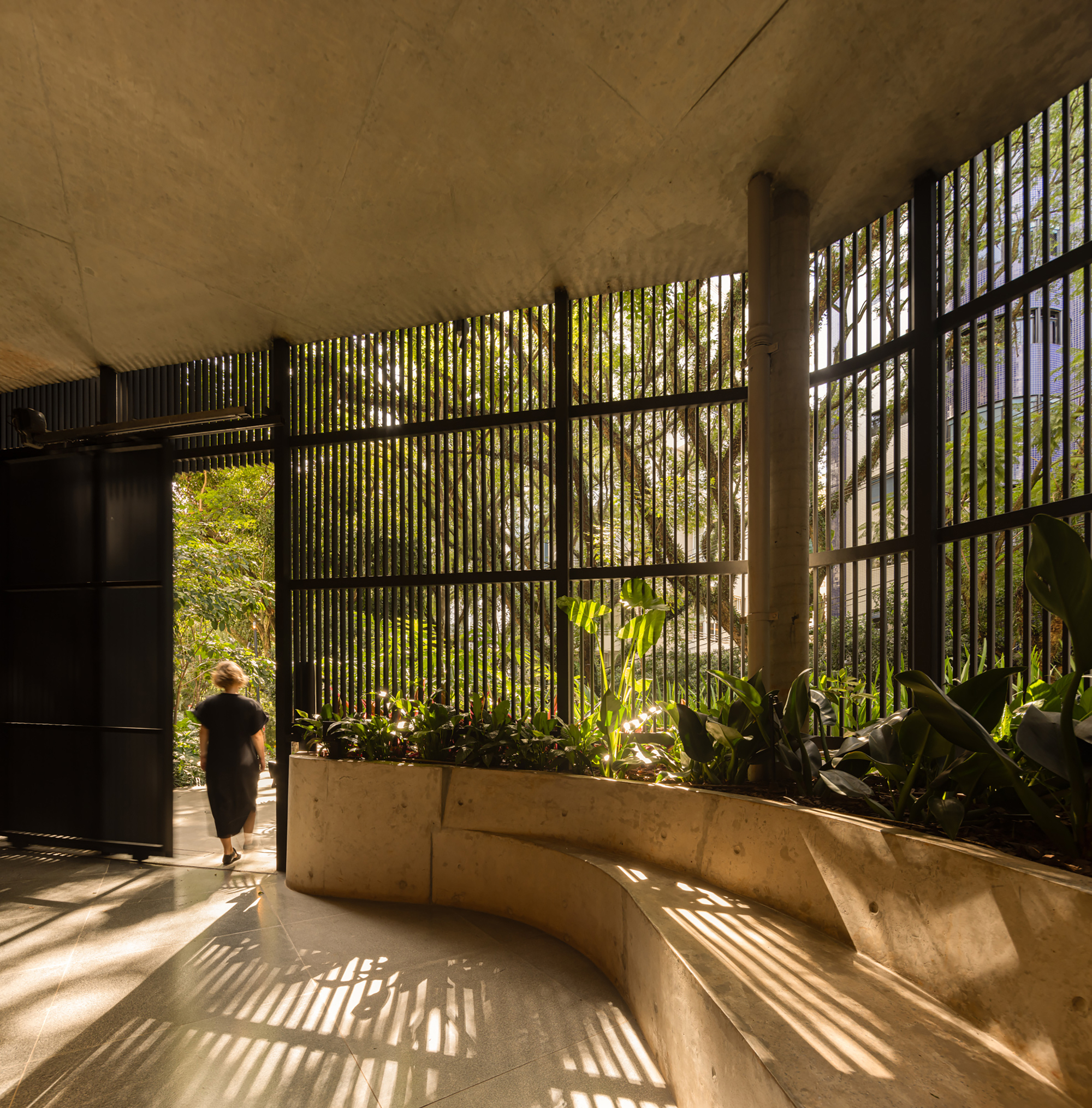 Lobby space with vertical brise-soleil casting striped shadows across curved concrete planter and stone floor