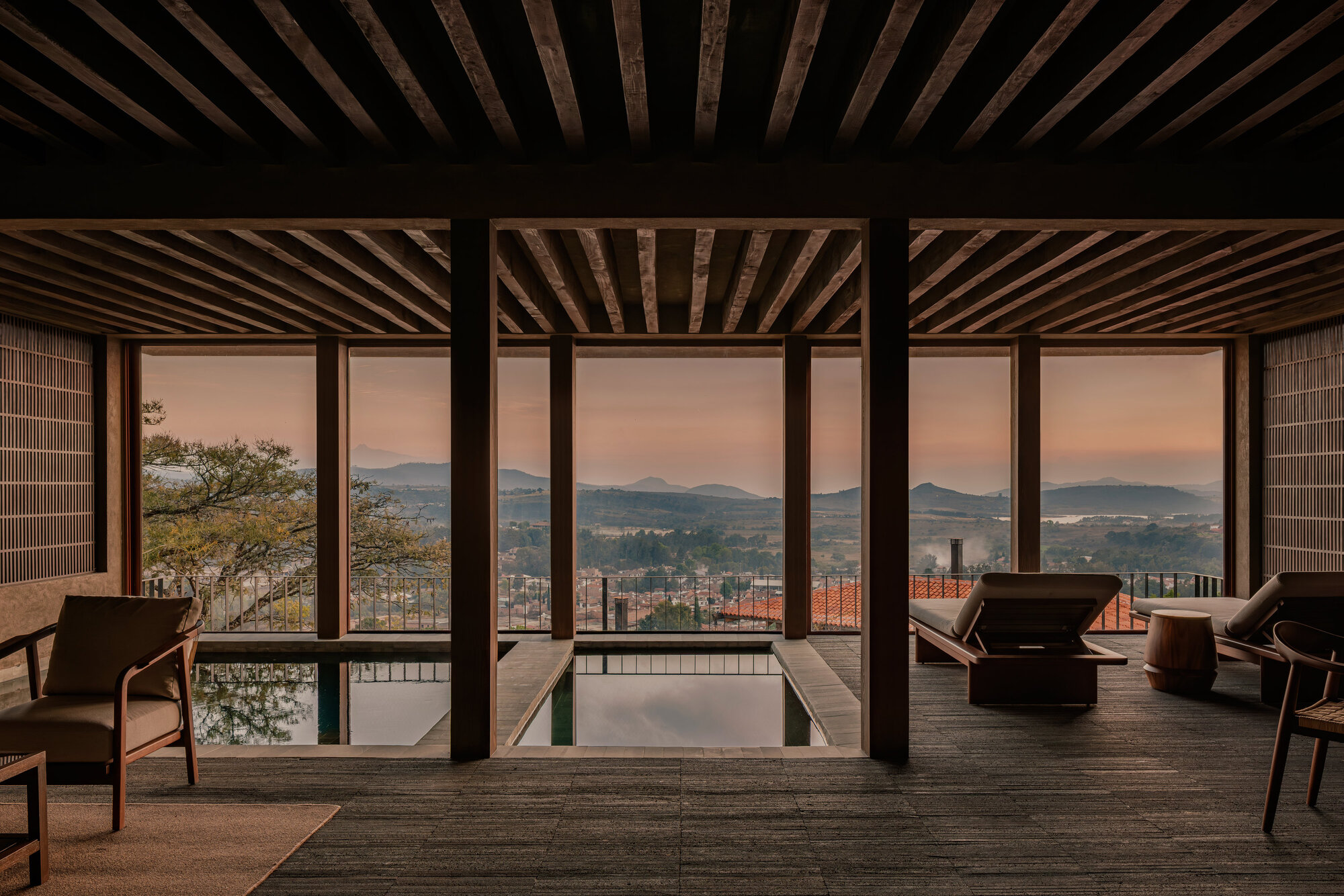 Interior pavilion with exposed timber ceiling beams and floor-to-ceiling glazing framing distant mountains at dusk
