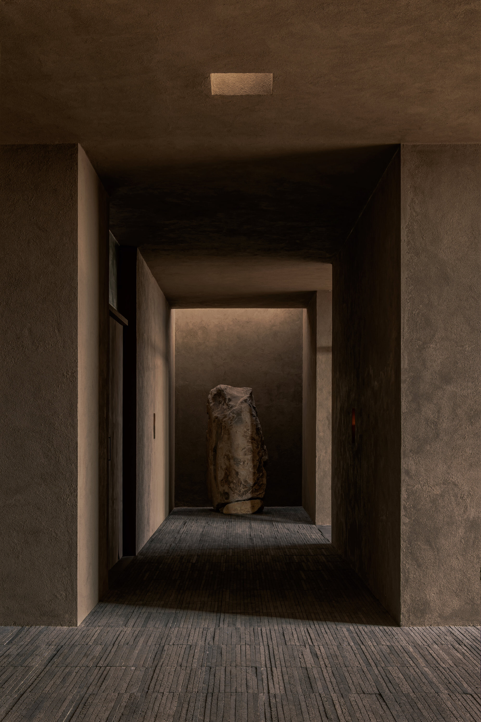 Compressed corridor sequence with layered thresholds framing a stone sculpture under recessed ceiling slot