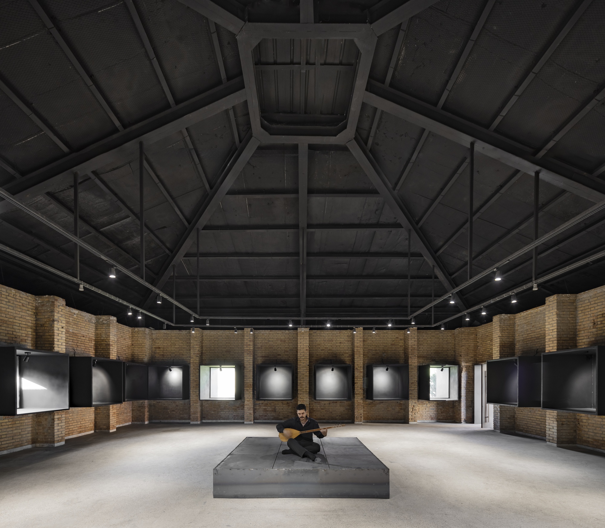 Interior gallery space with exposed timber roof beams radiating from a central skylight above a seated figure