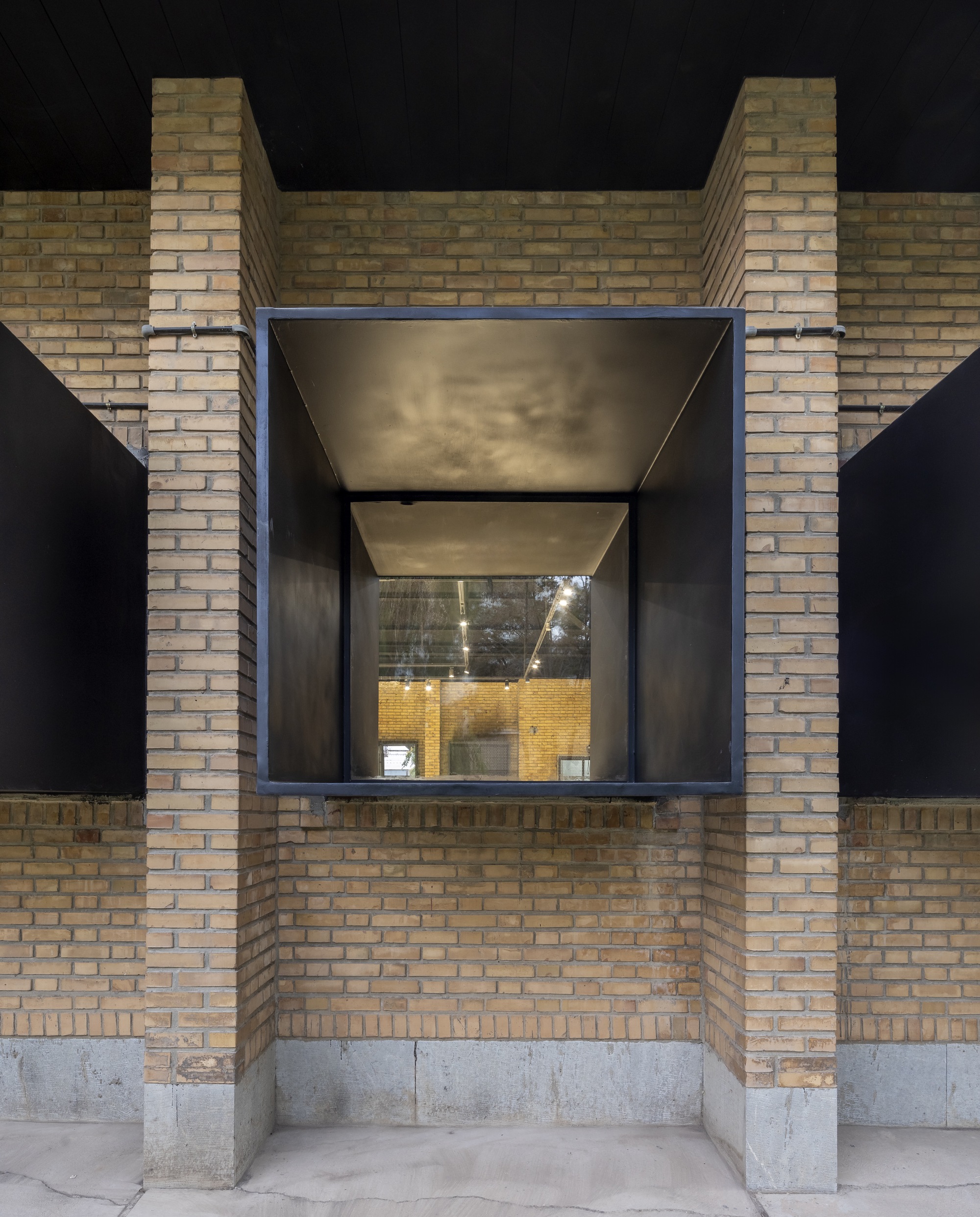 Recessed black steel window volume framed by yellow brick piers and concrete base at night