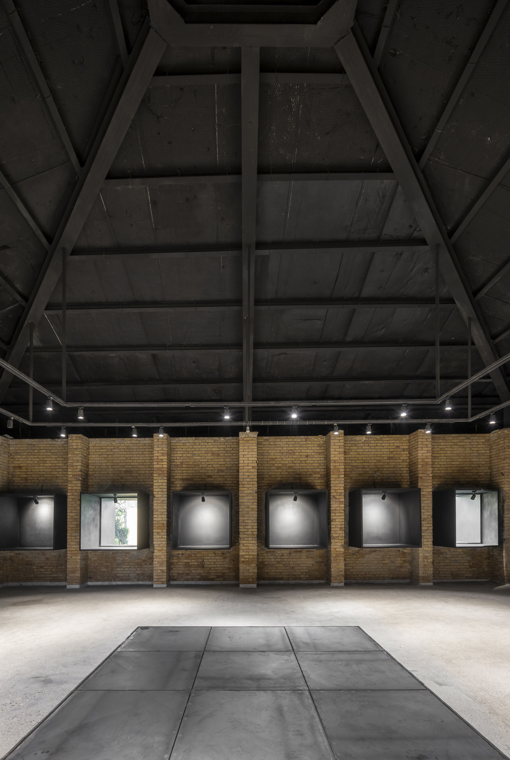 Interior gallery space with exposed timber roof trusses and framed artworks along yellow brick walls