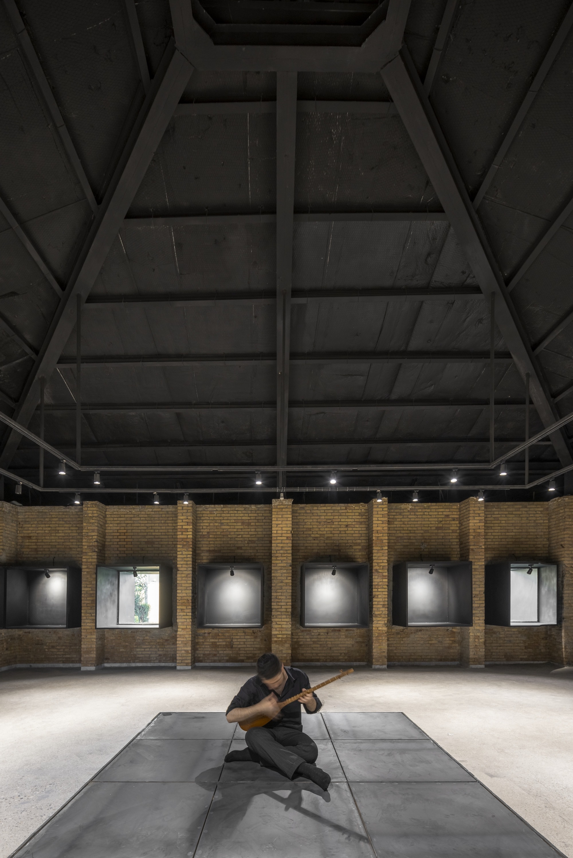 Gallery interior with a person seated on the central concrete platform beneath timber truss ceiling