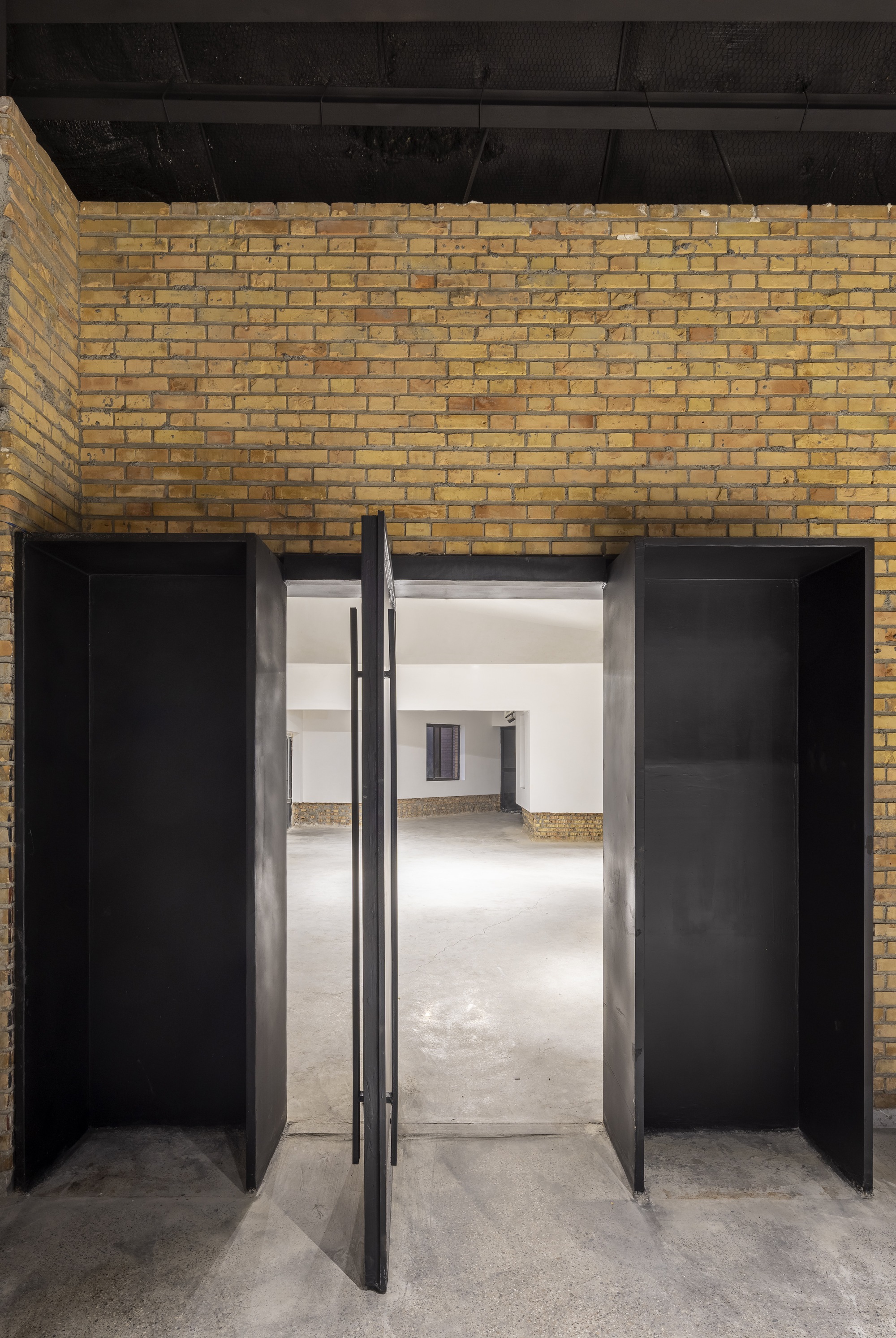 Pivot doors in black metal frames opening from a yellow brick wall into white-walled interior spaces
