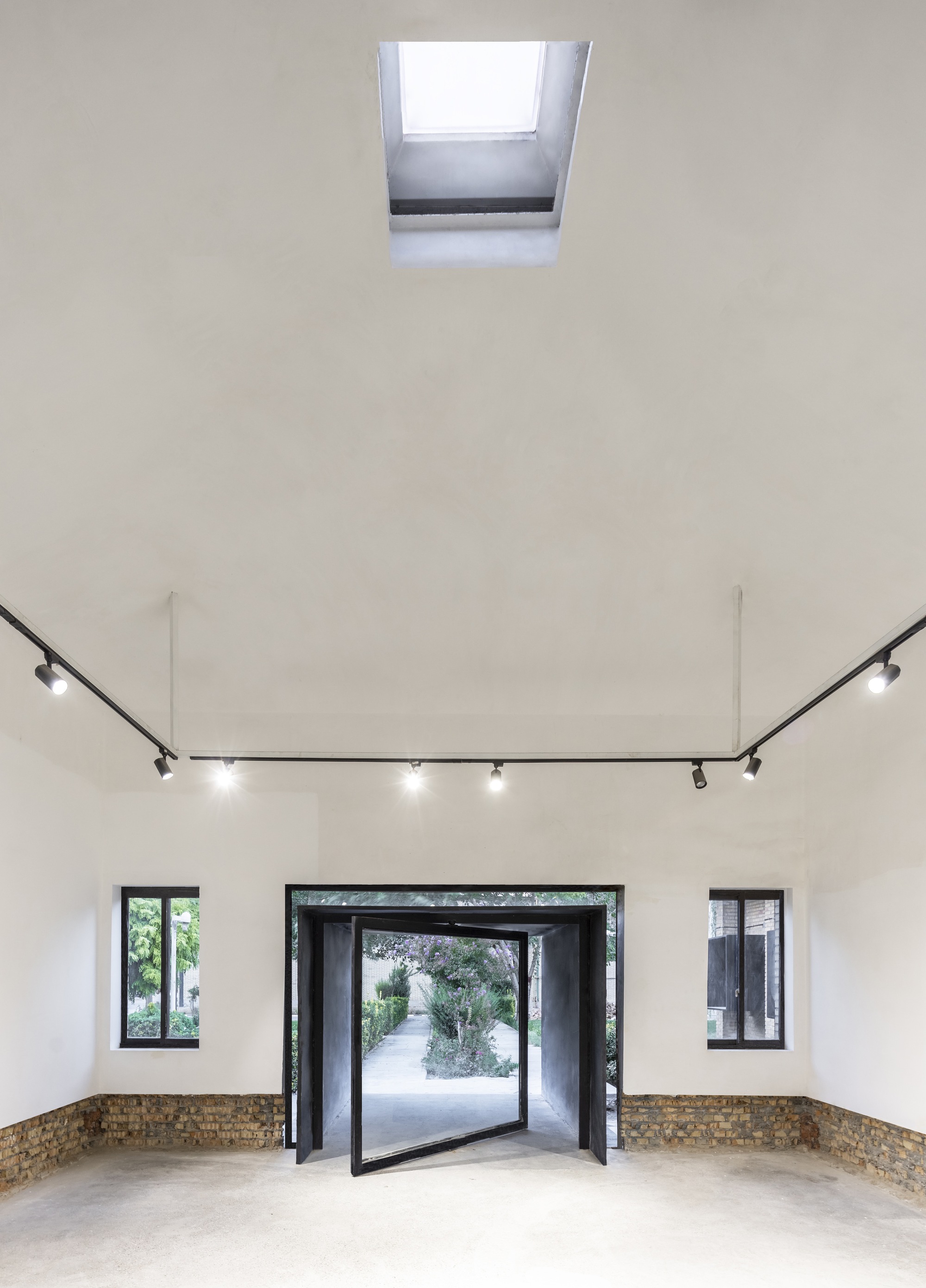 White-walled gallery with central pivot door, flanking windows, track lighting and a roof skylight above
