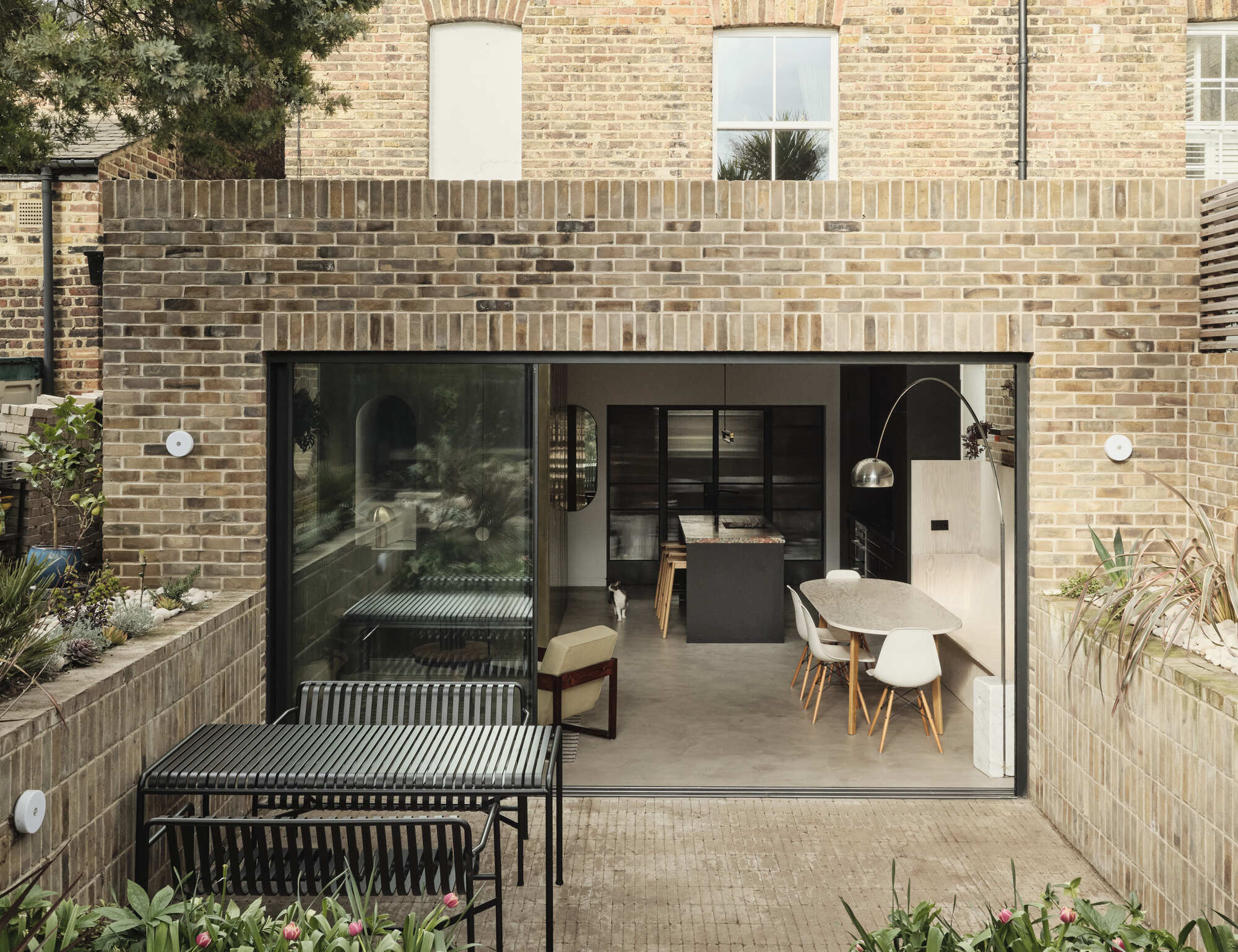 Rear courtyard with full-height glass doors opening to a kitchen and dining area beneath buff brick walls