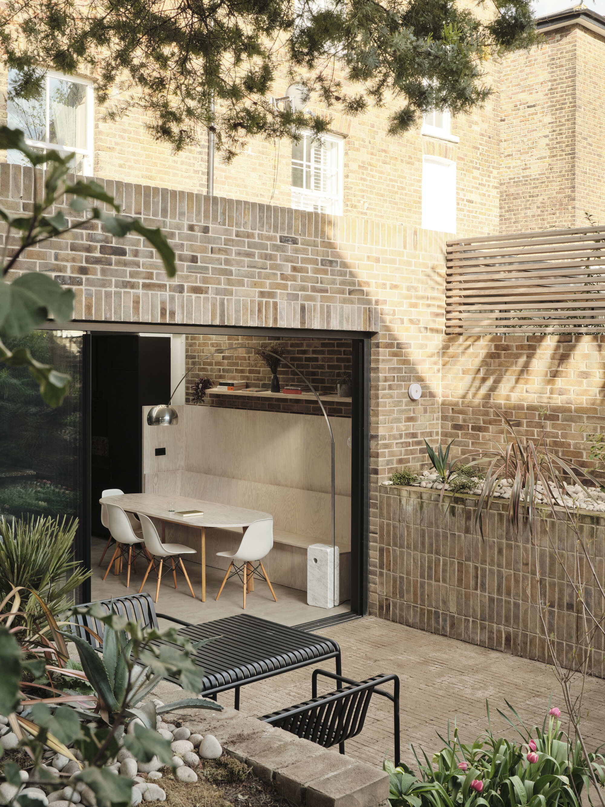 Glazed rear extension opening to courtyard garden with lounge chairs beneath overhanging tree branches