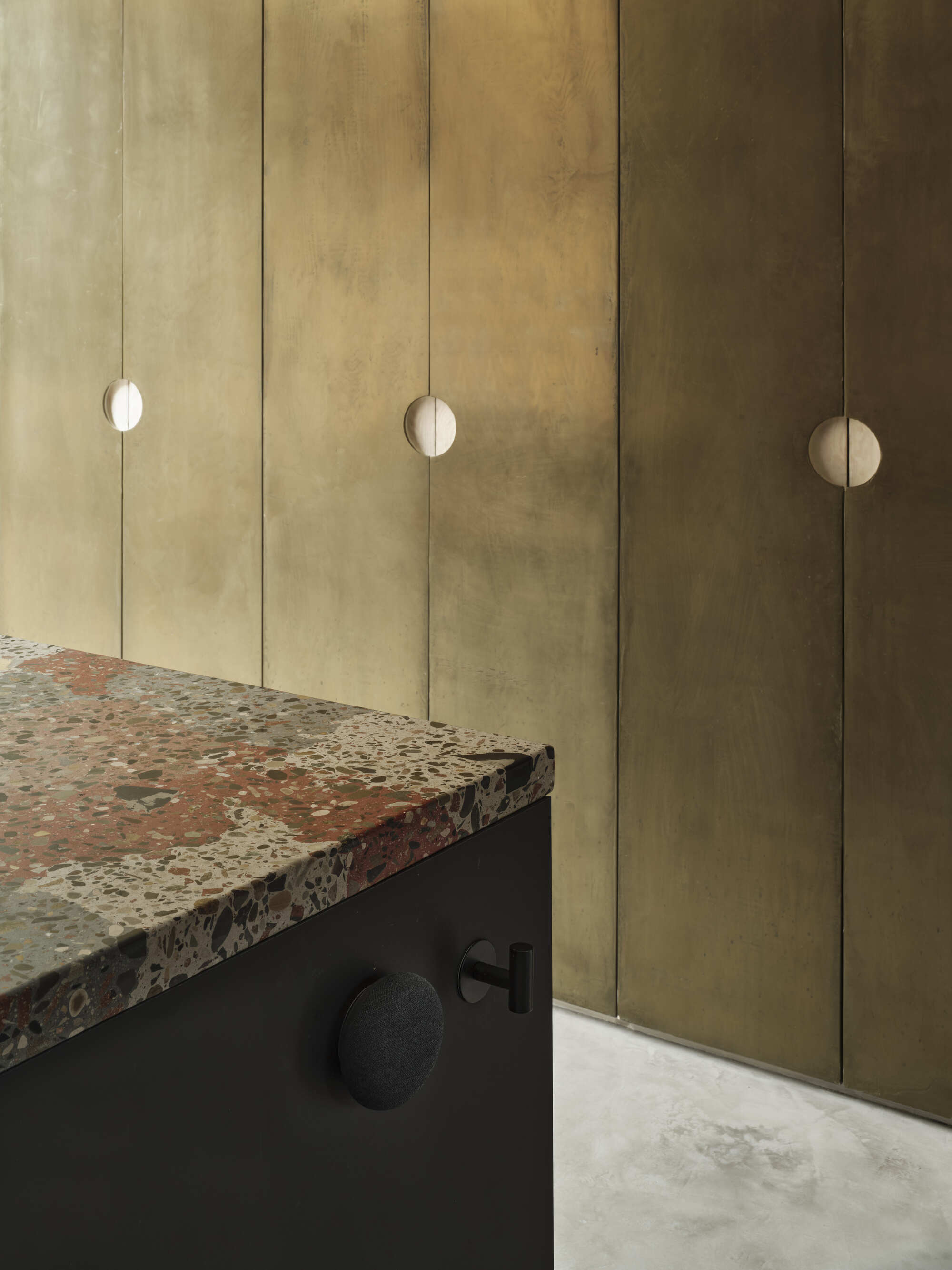 Brushed brass cabinet wall with circular recessed pulls beside a dark joinery unit with terrazzo countertop