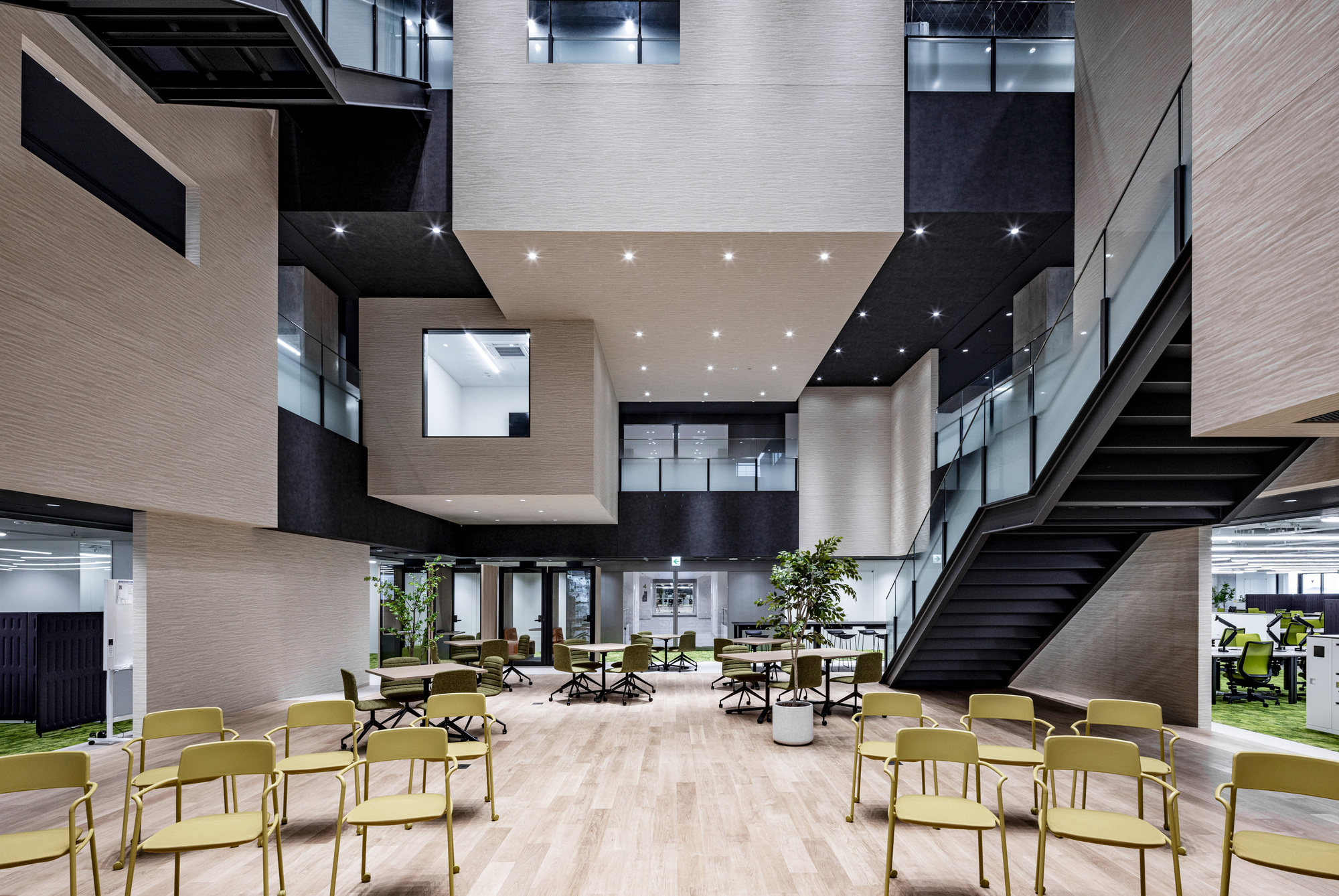 Multi-storey atrium with light wood-clad volumes, black ceilings and yellow chairs arranged on the ground floor
