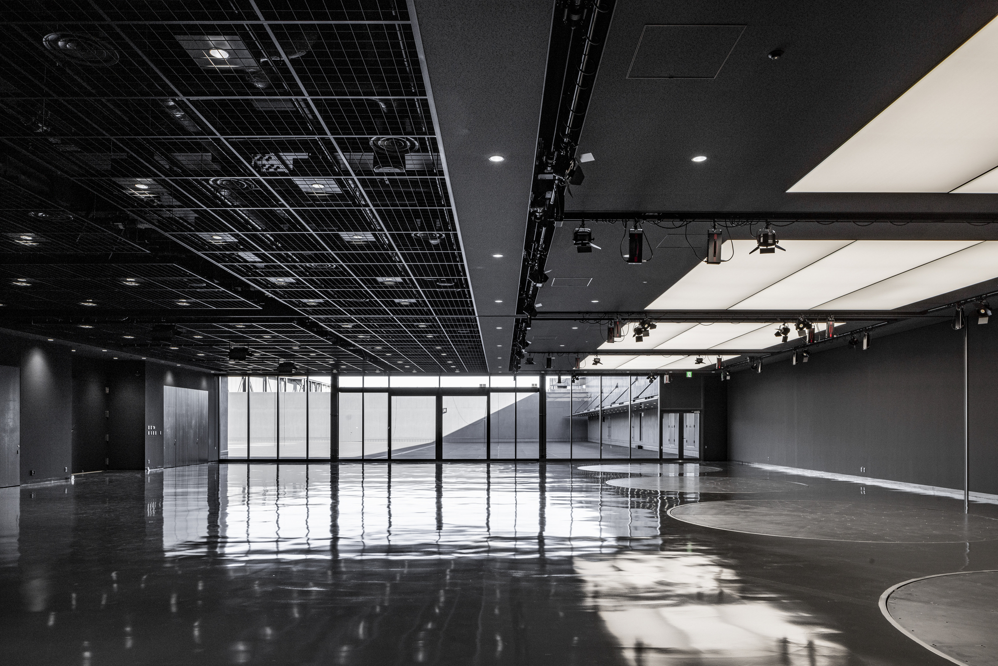 Open event space with black exposed ceiling grid and glazed walls overlooking an interior staircase