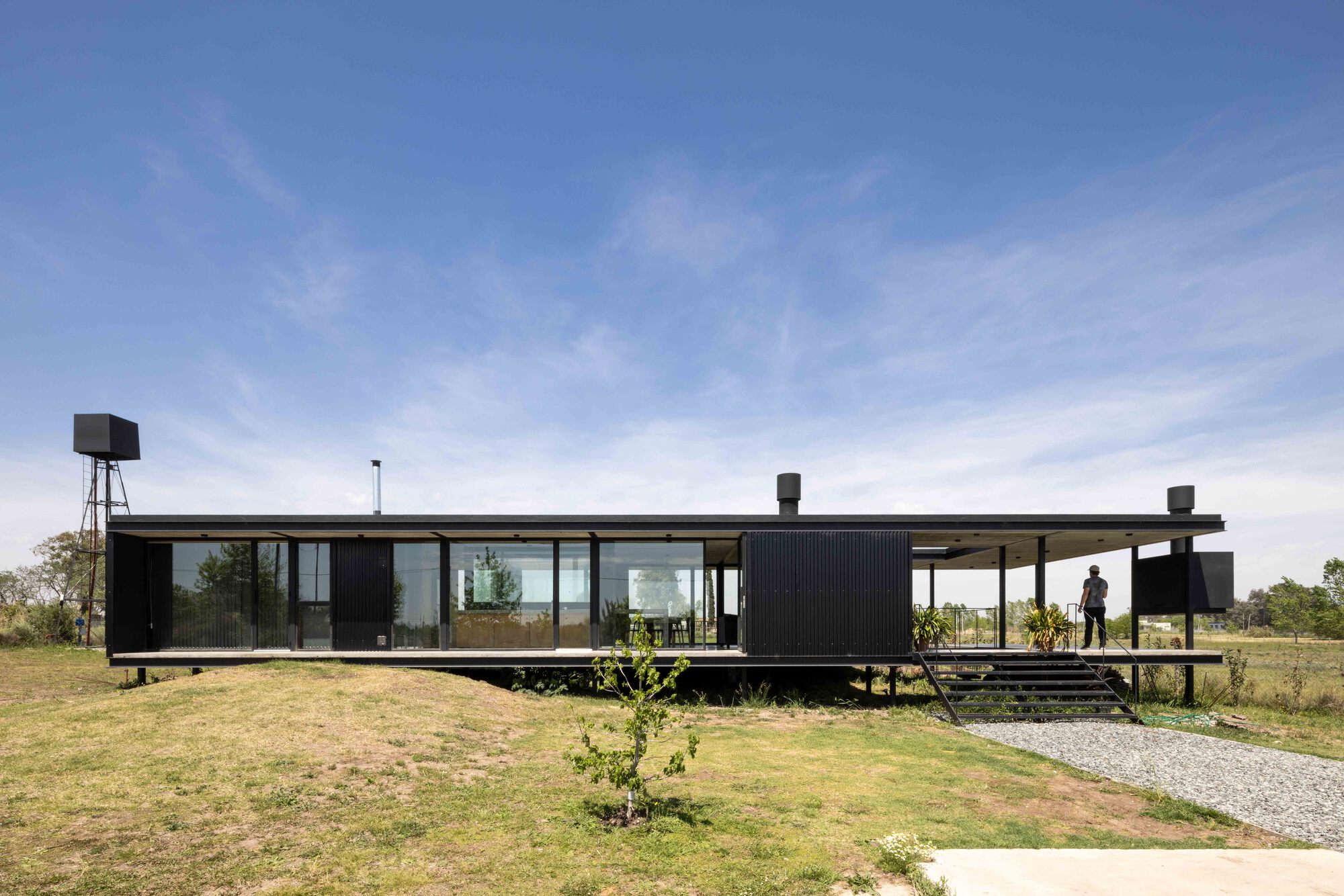 Elevated black corrugated metal pavilion with full-height glazing and covered terrace in open grassland
