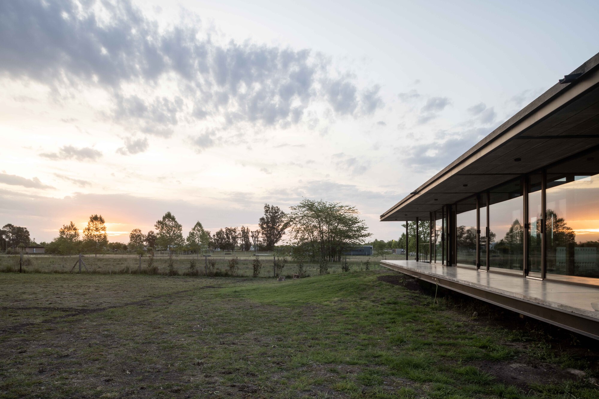 Elevated glass pavilion on a raised platform overlooking open grassland at sunset