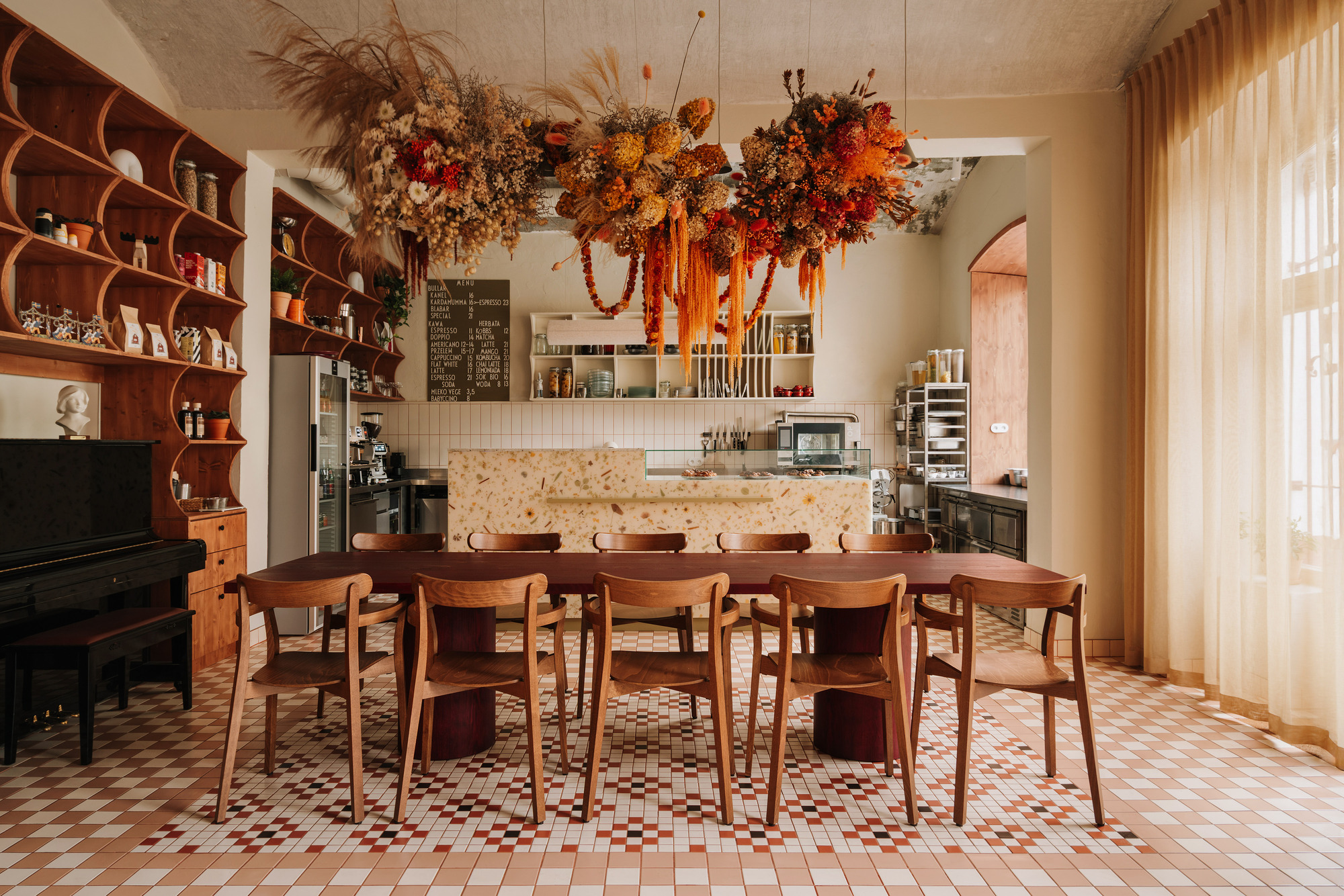 Long timber communal table beneath large suspended dried floral arrangement in room with chequered floor