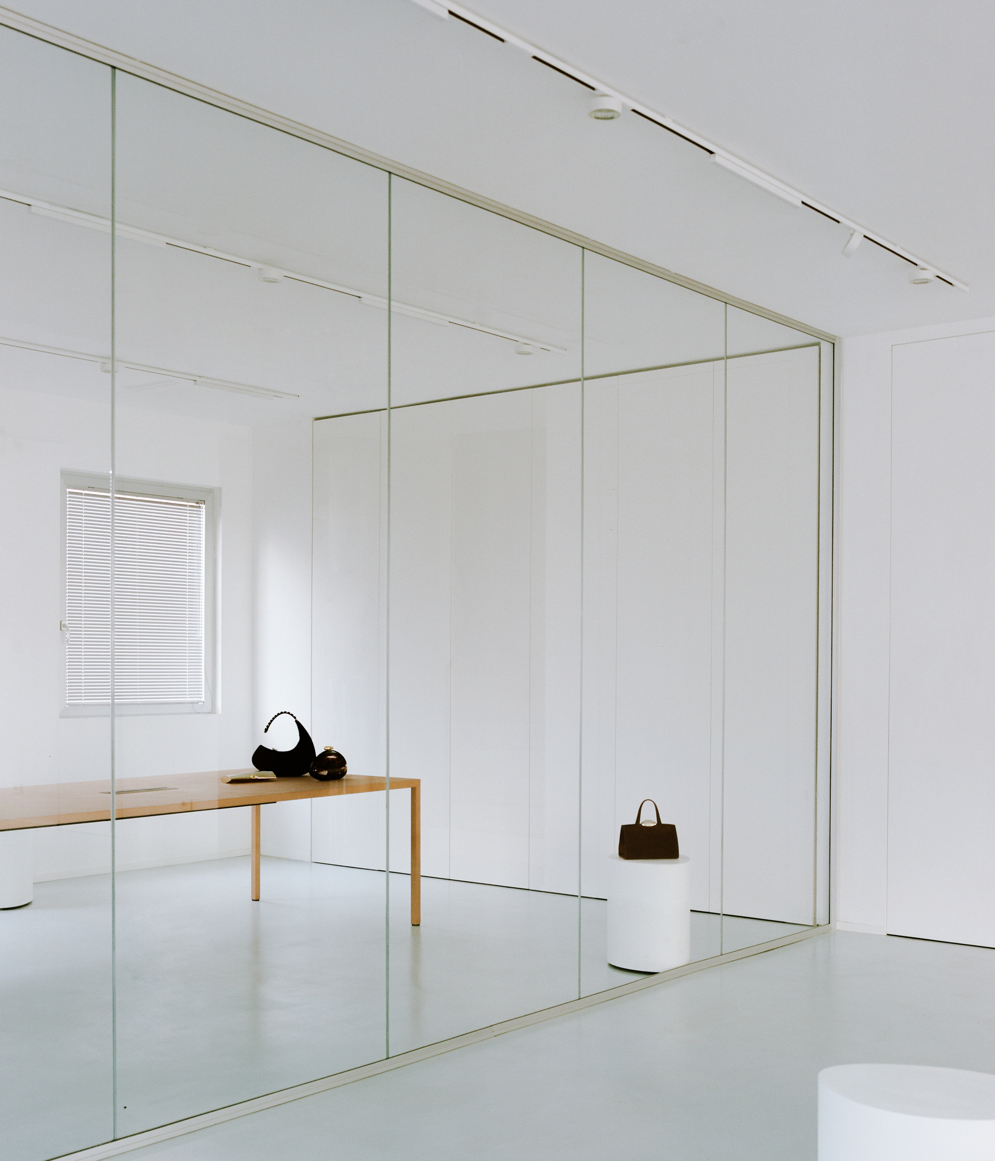 Floor-to-ceiling glass partition reflecting a timber bench and handbags with track lighting on the ceiling