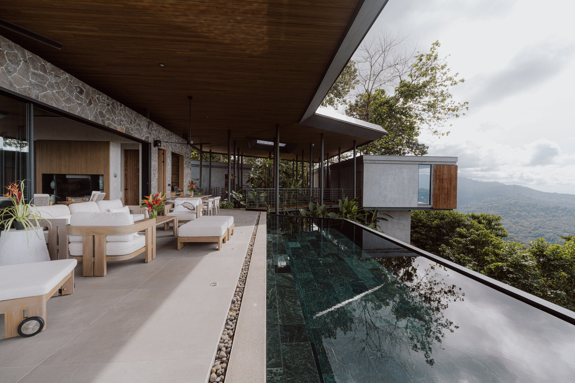 Covered terrace with infinity pool, timber ceiling, stone wall, and mountain vista beyond