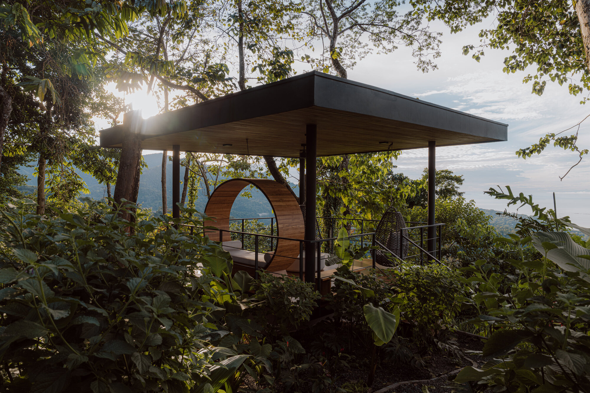 Flat-roofed pavilion with circular timber portal framing mountain views among tropical vegetation at sunset