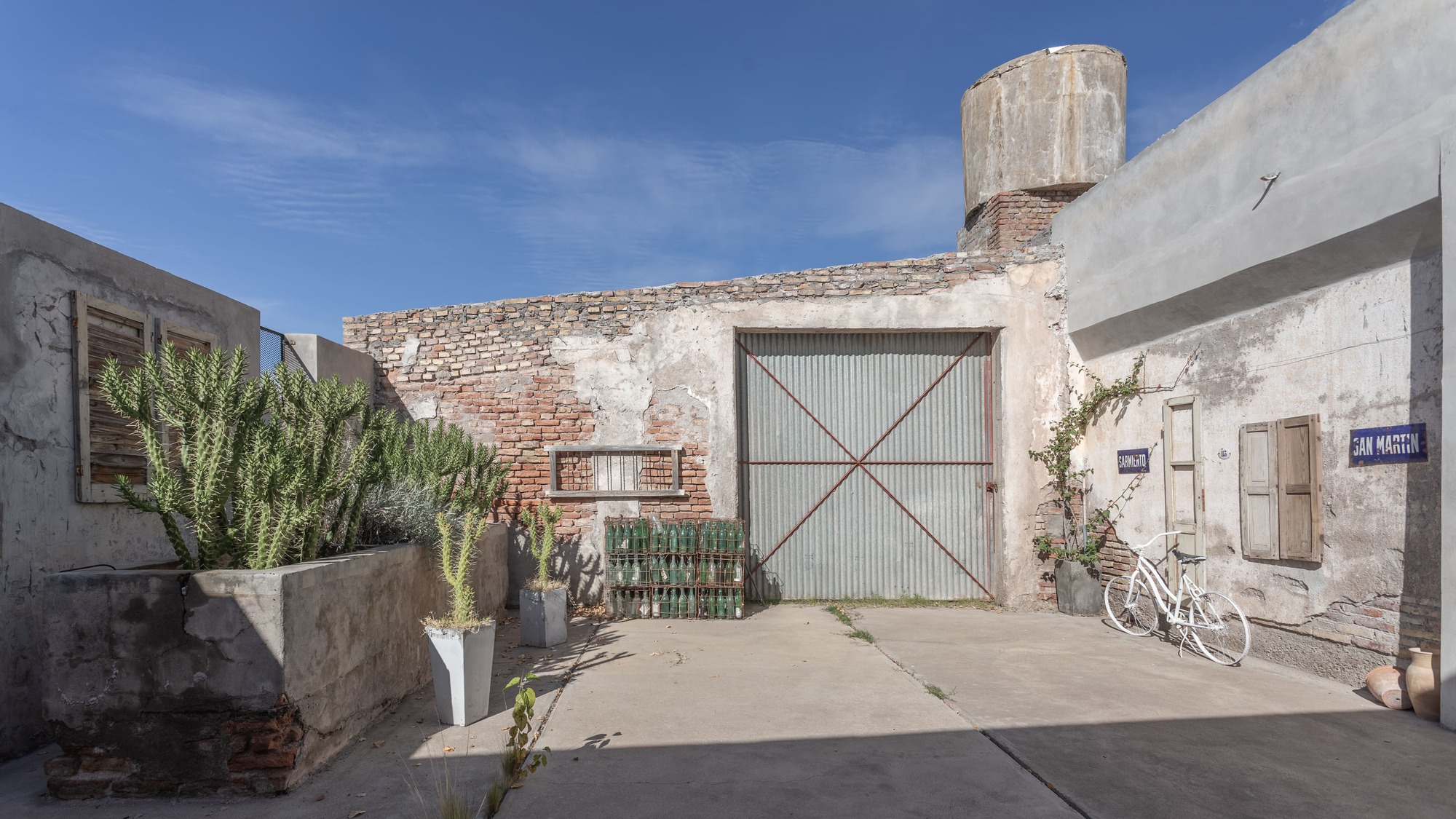 Courtyard entry with corrugated metal gate flanked by planted cacti and a bicycle under clear skies