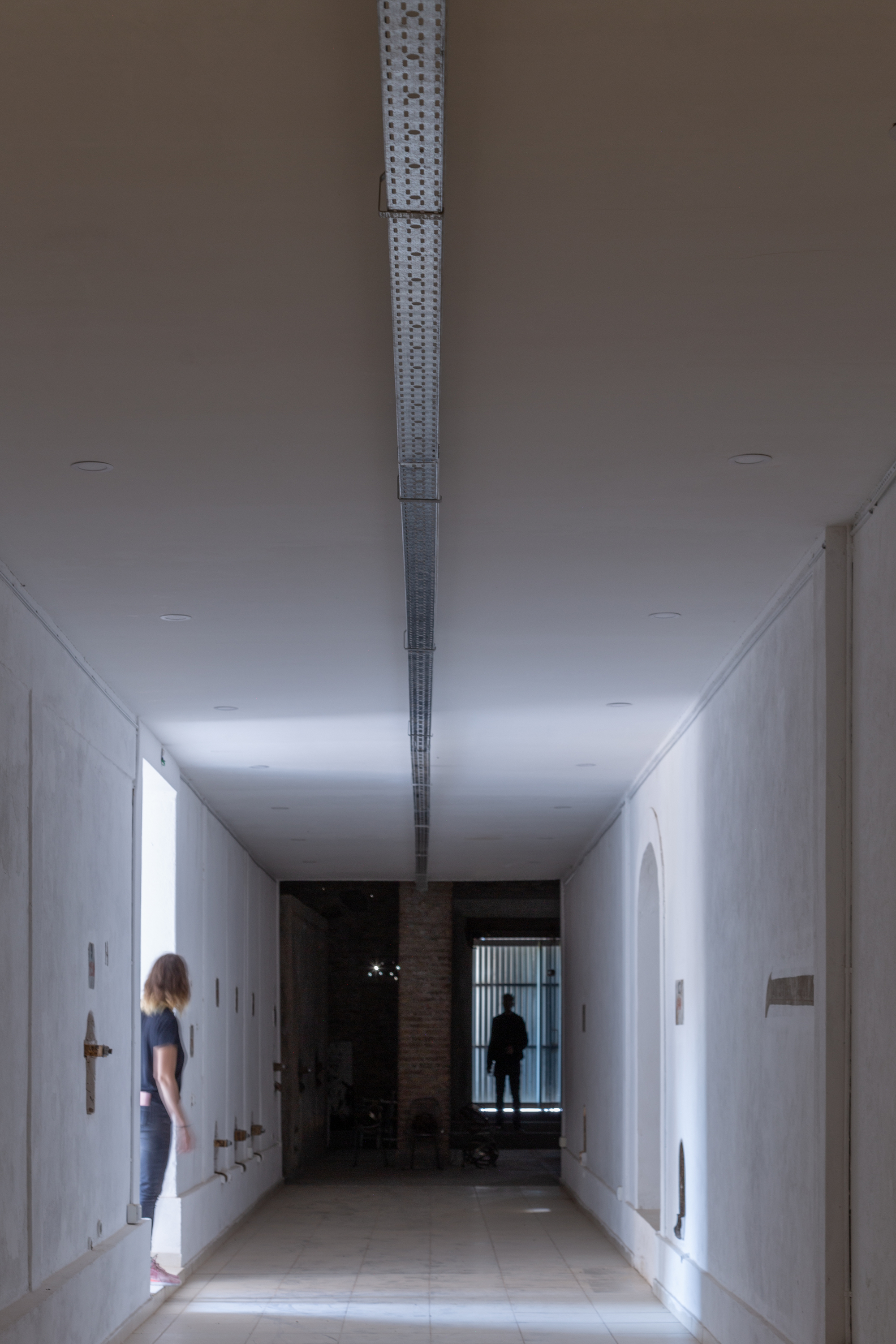 Long white corridor with perforated metal skylight running along the ceiling center and two figures