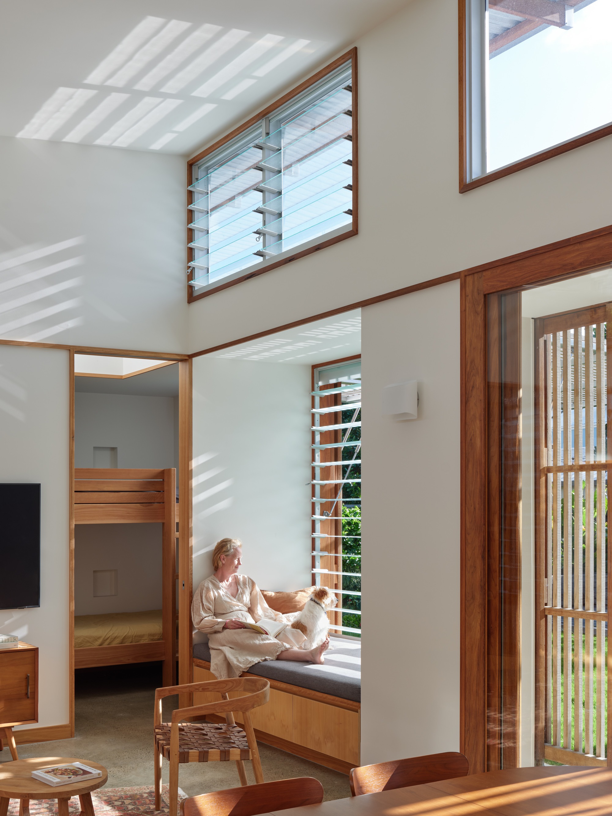 Double-height living area with woman seated on window bench beneath louvered windows in afternoon light