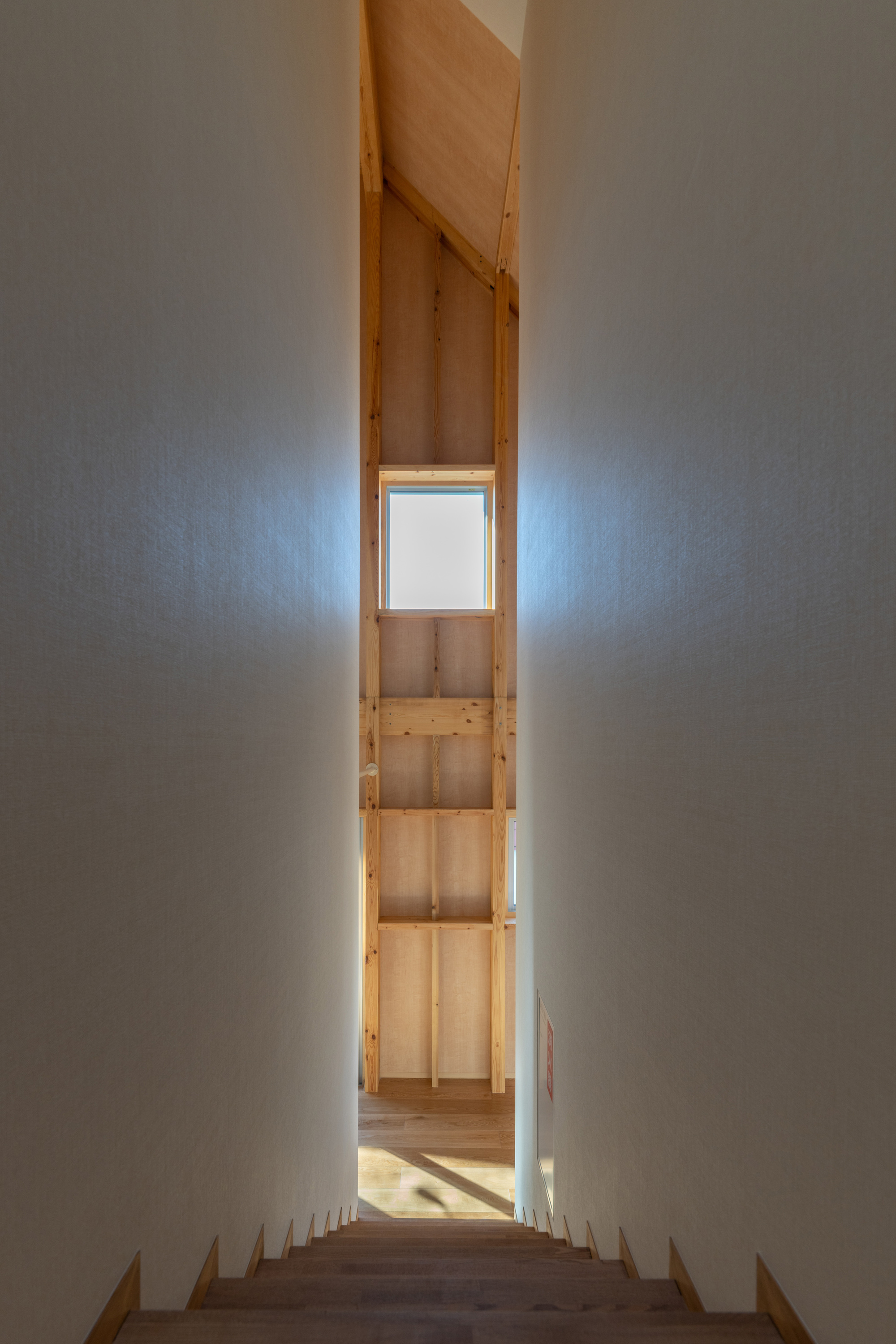 View down a timber staircase toward a narrow landing with a square window framing daylight