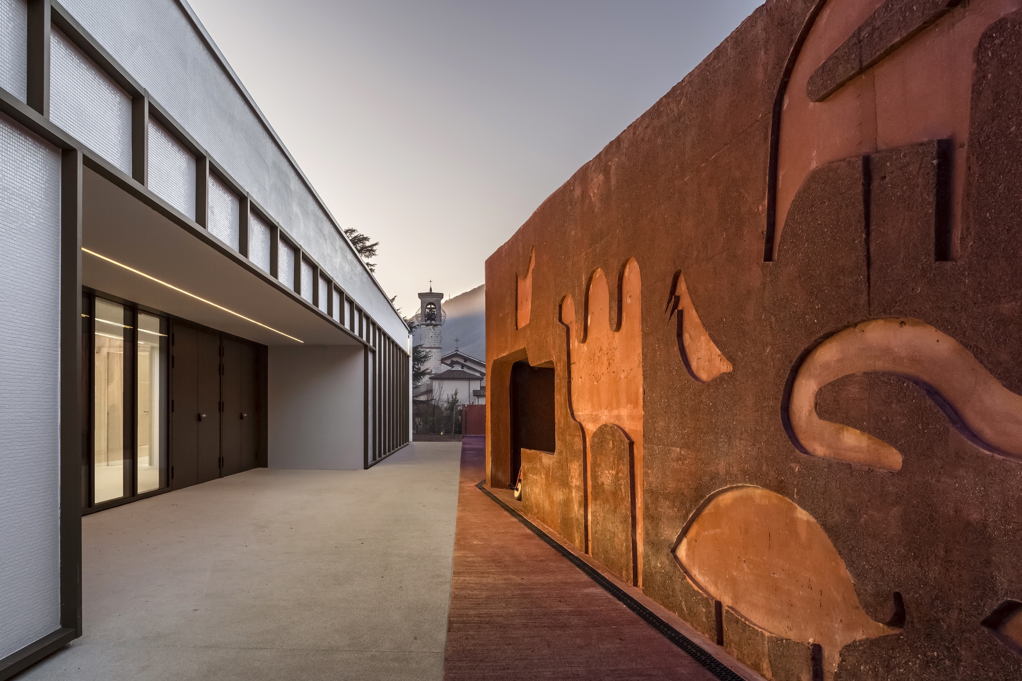 Courtyard passage between weathered steel sculptural wall and white volume with vertical glazing at dusk