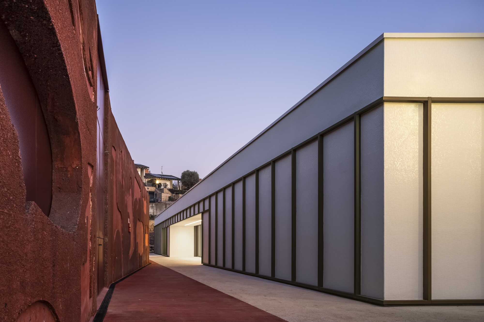 Narrow courtyard between the rammed earth wall and the translucent panel facade at dusk