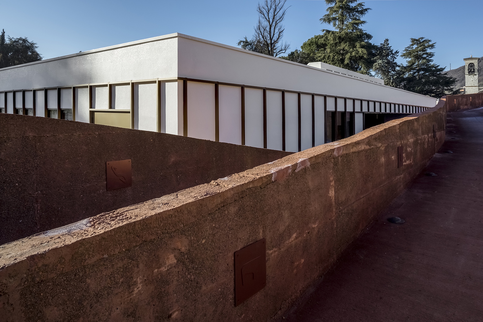 Long view of the white panel facade above the weathered rammed earth wall in daylight