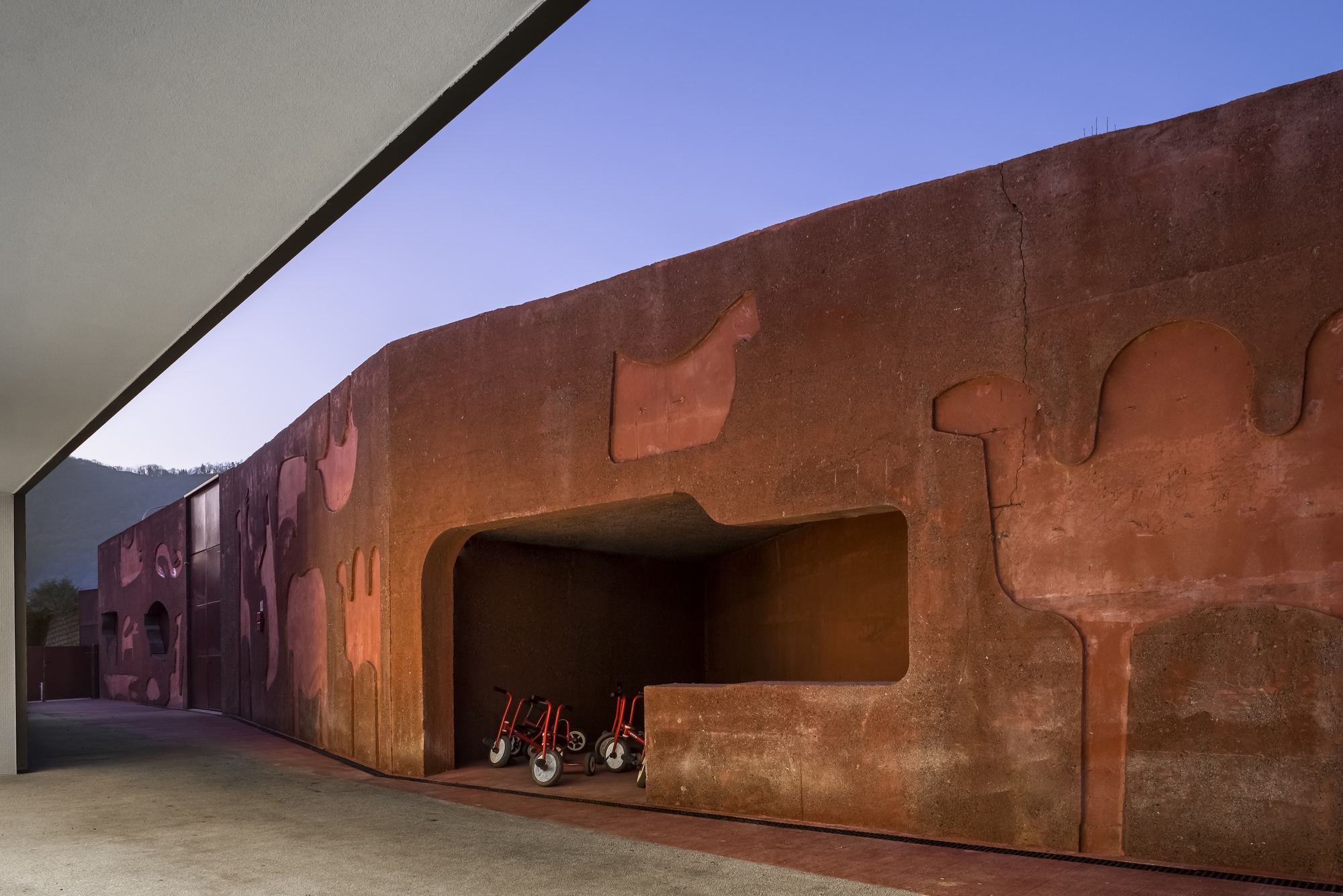 Rammed earth wall with sculpted relief cutouts and wheelchairs stored in a recessed niche